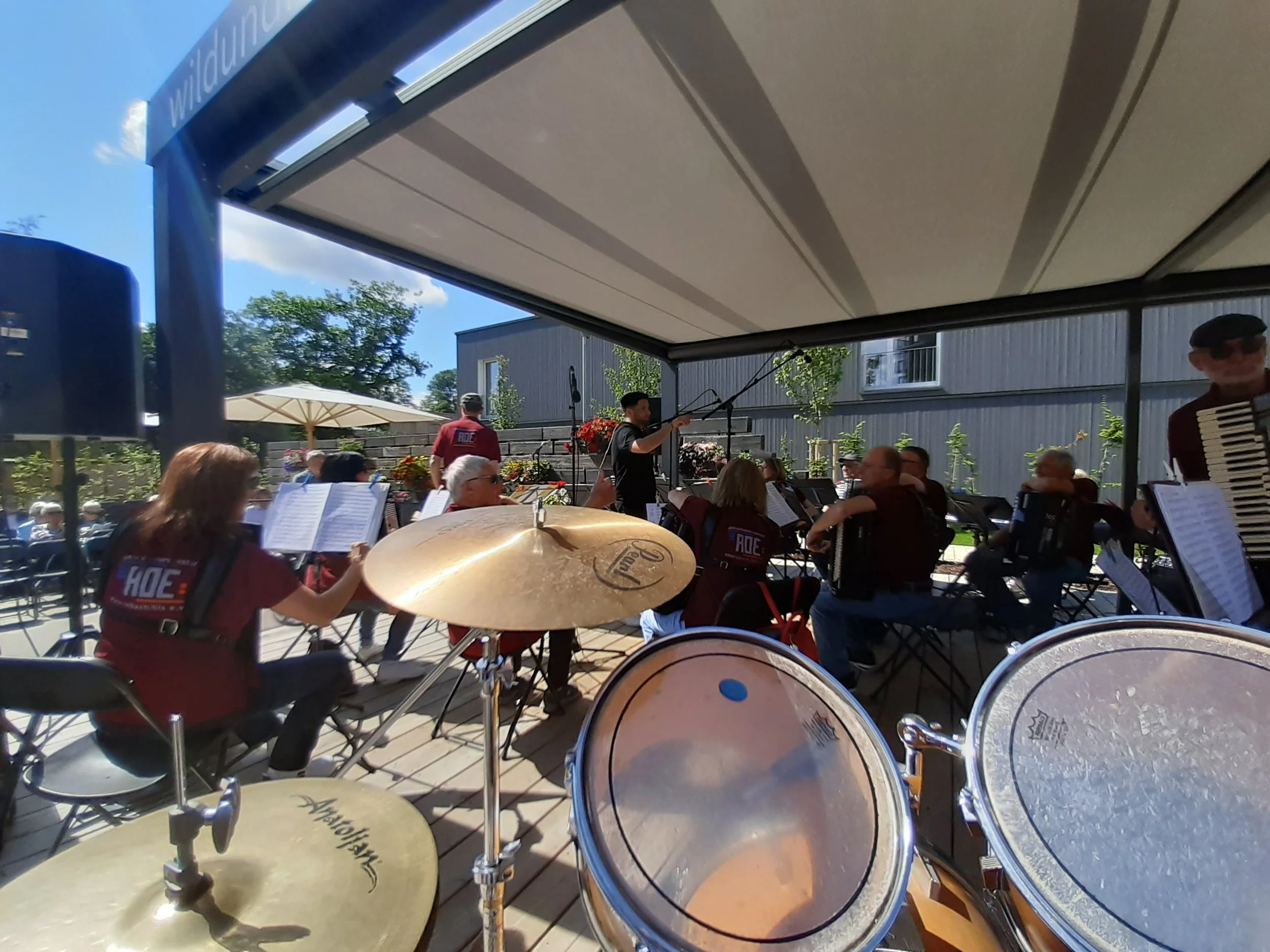 Blick auf eine Outdoor-Konzerthalle mit einer Bigband, bei der Musiker mit Instrumenten wie Trommeln und Akkordeons sitzen. Im Hintergrund sind Musiker auf einer Bühne mit Mikrofonen und illustrative Pflanzen sichtbar.