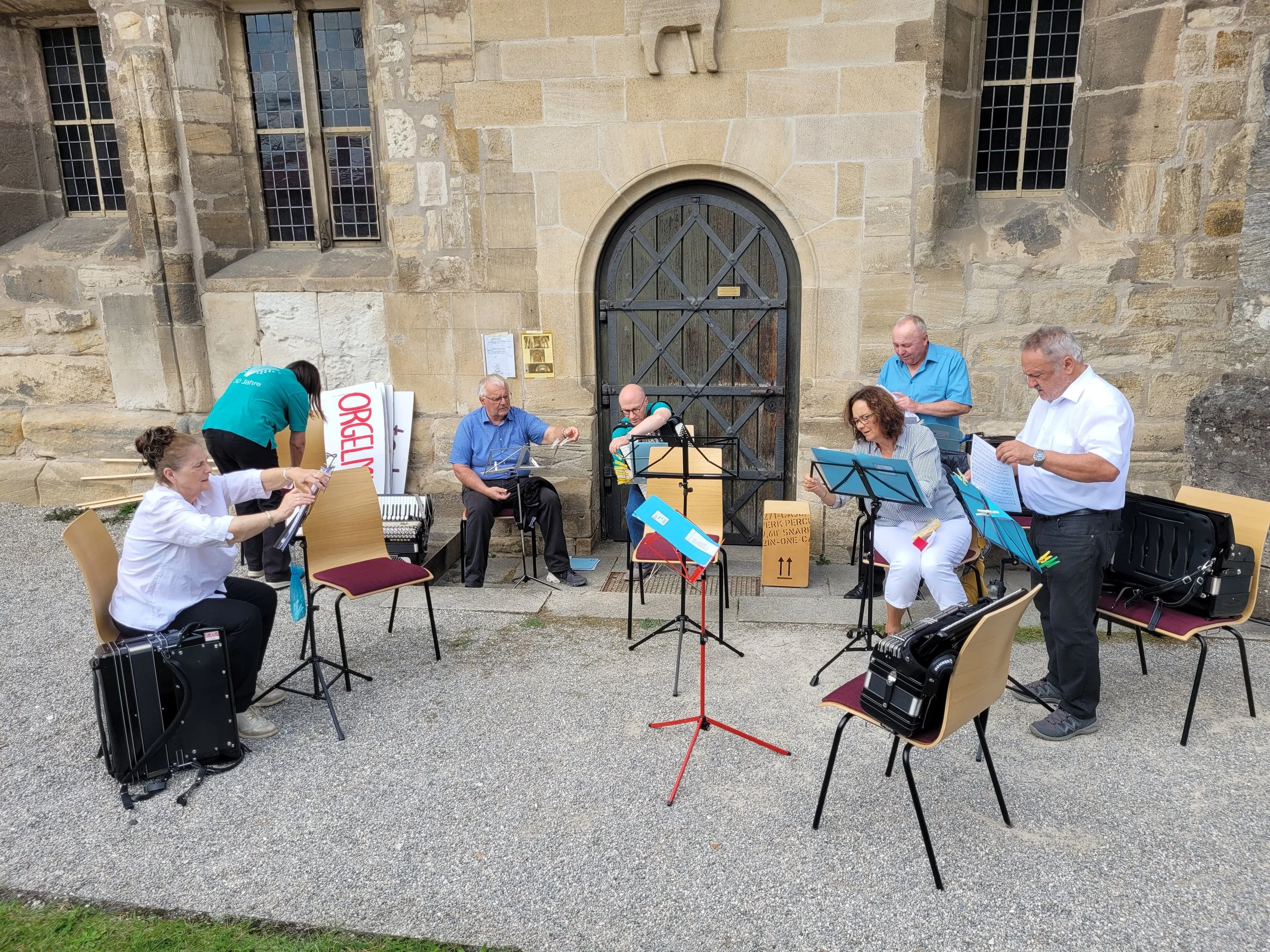 Eine Gruppe von Musikern spielt draußen vor einer alten Steingemeinde mit einem großen Holztor. Es sind Männer und Frauen, die verschiedene Instrumente spielen, darunter Klavier, Gitarre, Flöte und andere. Einige sind vertieft in ihre Musik, während andere lesen oder zuhören.
