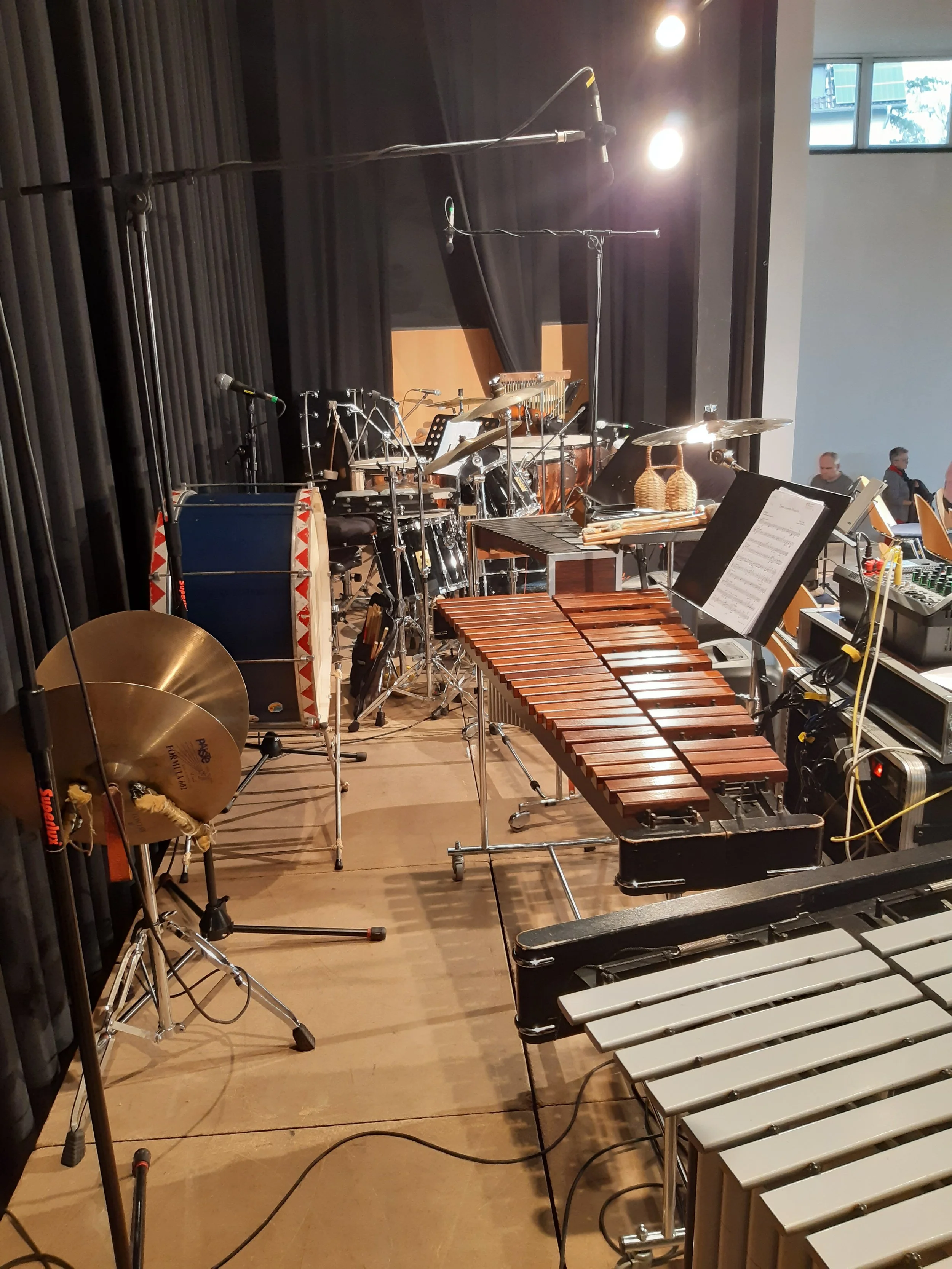 Blick auf eine Bühne mit Musikinstrumenten, darunter Trommel, Xylophon, Marimbas, Becken, Gongs, Musiknotenständer, Mikrofone, Kabel und Lichtquellen.