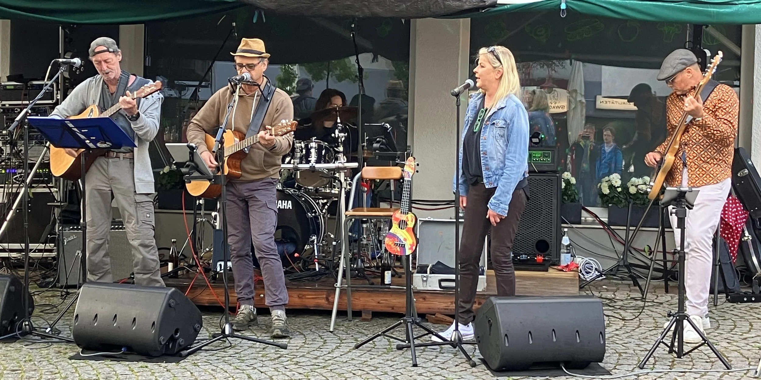 Eine Band spielt auf einer kleinen Open-Air-Bühne, bestehend aus mehreren Musikern mit Gitarren und Schlagzeug, vor einem Publikum.