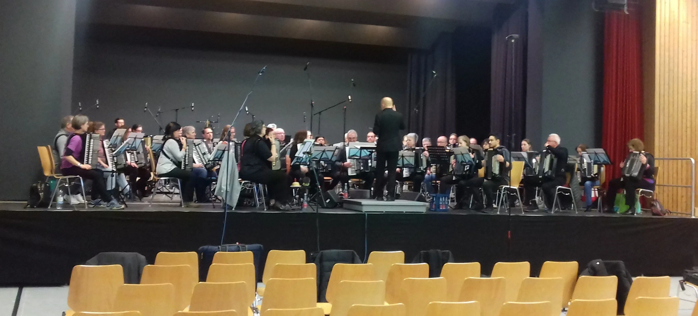 Ein Orchester spielt auf einer Bühne, mit einem Dirigenten in schwarzer Kleidung, umgeben von Musikern, die Akkordeons spielen. Es sind Mikros und Notenständer sichtbar.