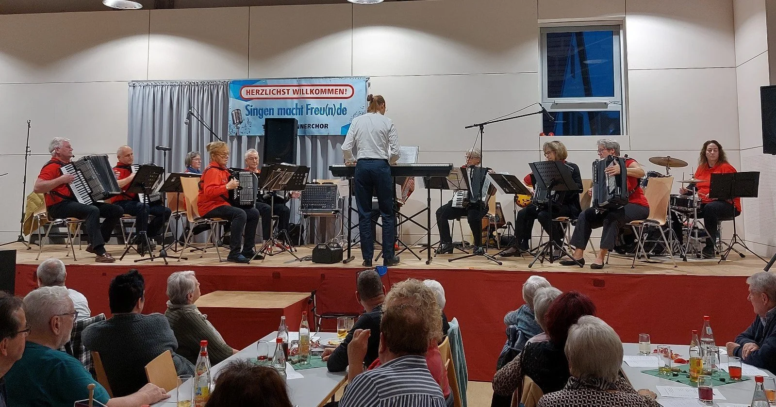 Eine Gruppe von Musikerinnen und Musikern spielt auf einer Bühne, während ein Dirigent die Leitung übernimmt. Das Publikum sitzt an Tischen und schaut zur Bühne.