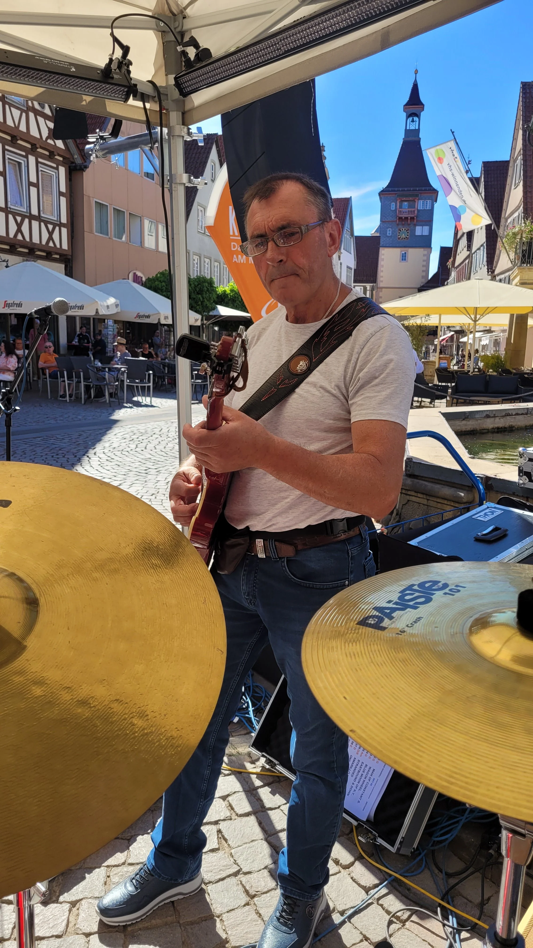 Mann spielt Gitarre während eines Live-Auftritts im Freien auf einer Terrasse vor historischen Gebäuden und einem Kirchturm.