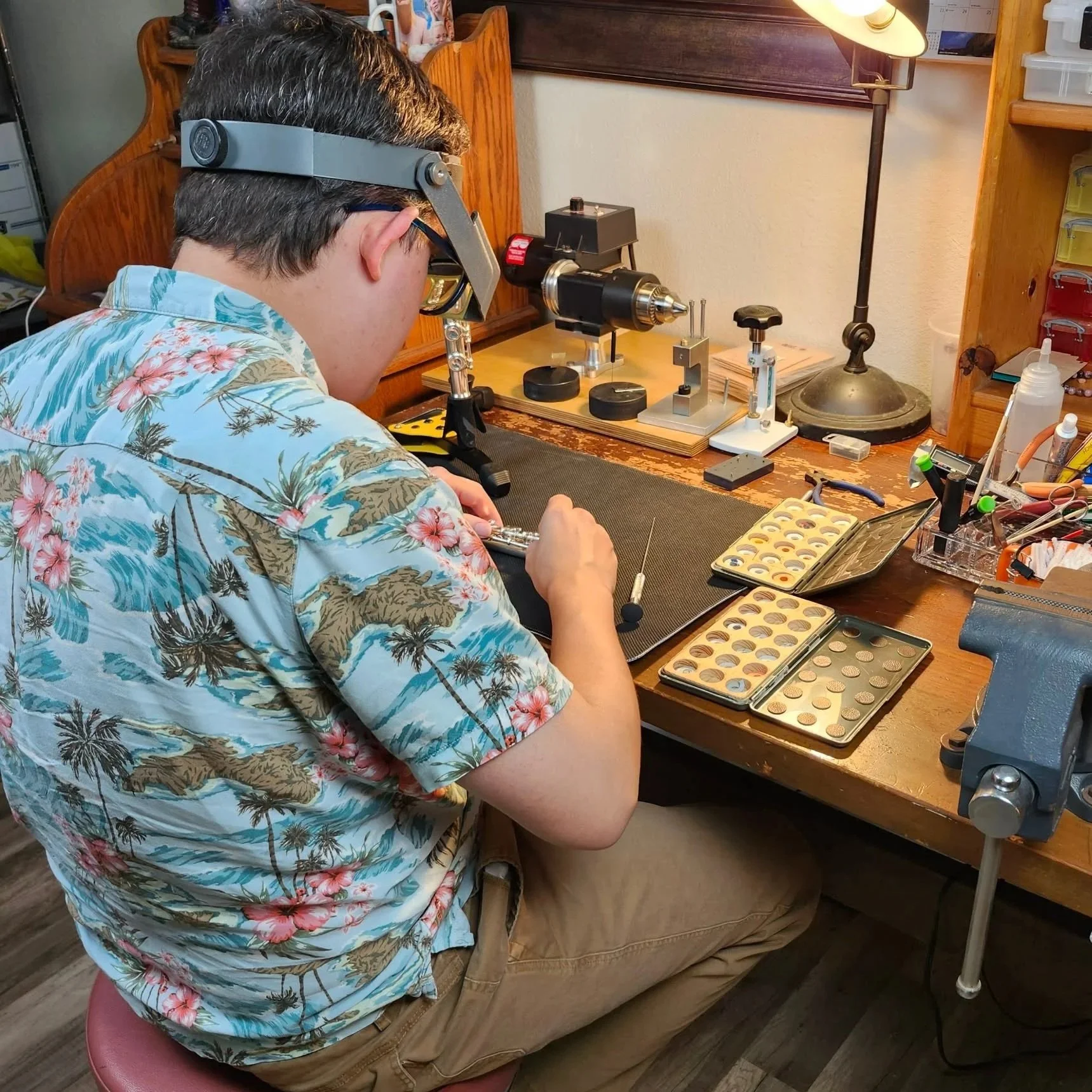 A person wearing glasses and a blue Hawaiian shirt with floral and palm tree patterns, sitting on a pink stool. They are working with various small objects on a wooden desk, which has multiple trays filled with circular items, tools, and equipment related to watch or jewelry repair. There is a lamp, a black work vise, and organized drawers nearby.