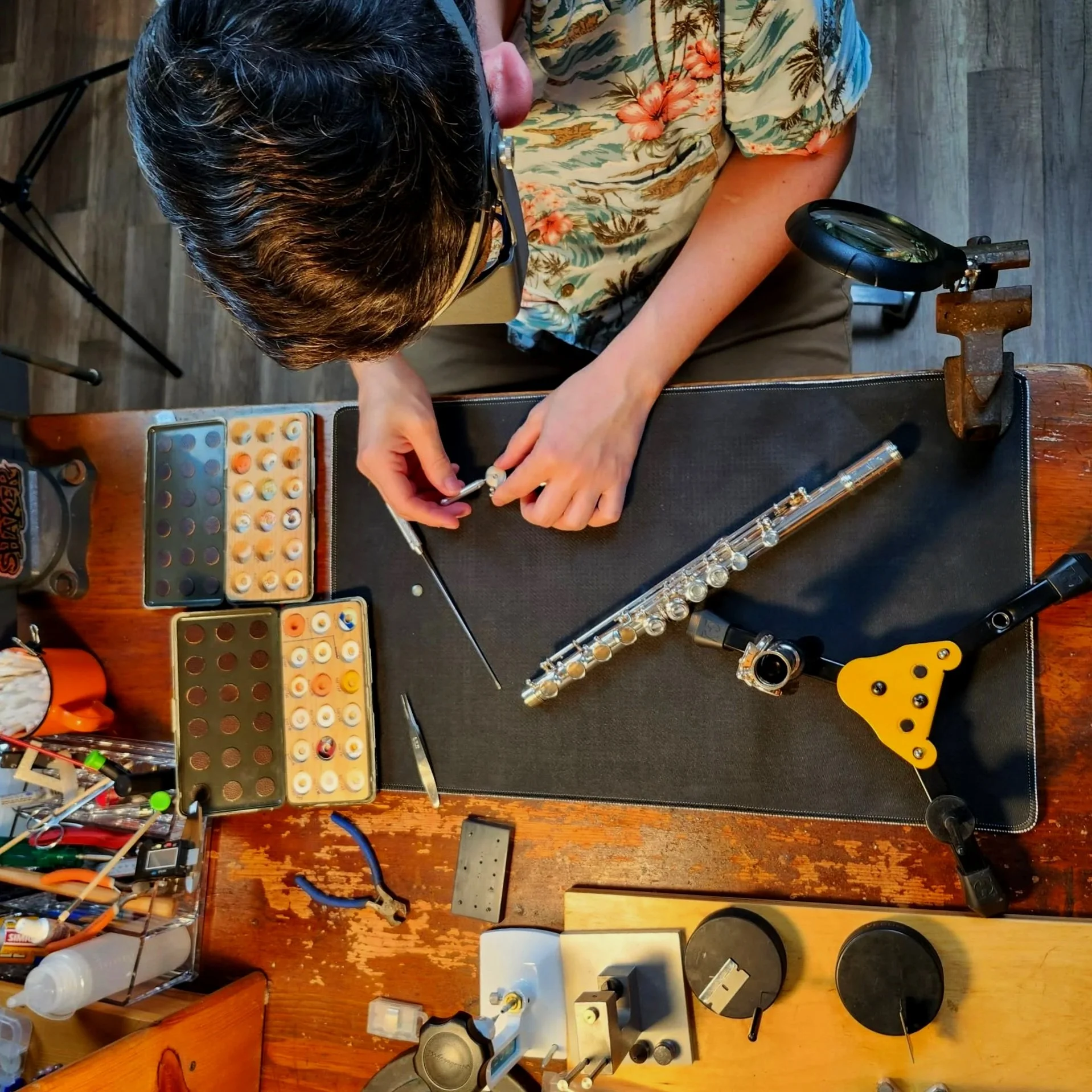 Top-down view of a person in a floral Hawaiian shirt working at a cluttered desk with music and electronics equipment, including a flute, compact discs, and electronic parts.