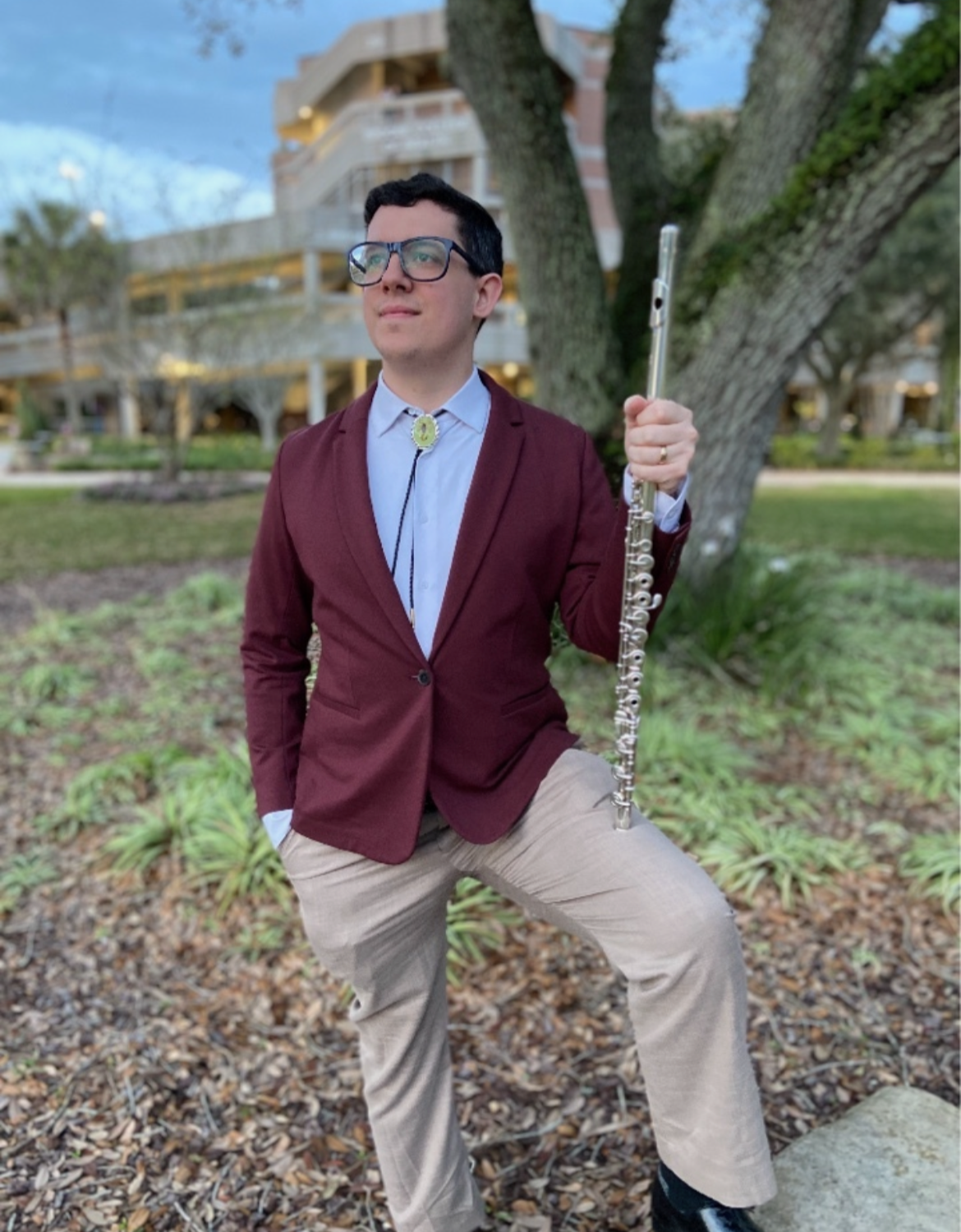 A man standing outdoors in front of a large tree, wearing glasses, a maroon blazer, white shirt, beige pants, holding a silver flute, with a modern apartment building in the background.
