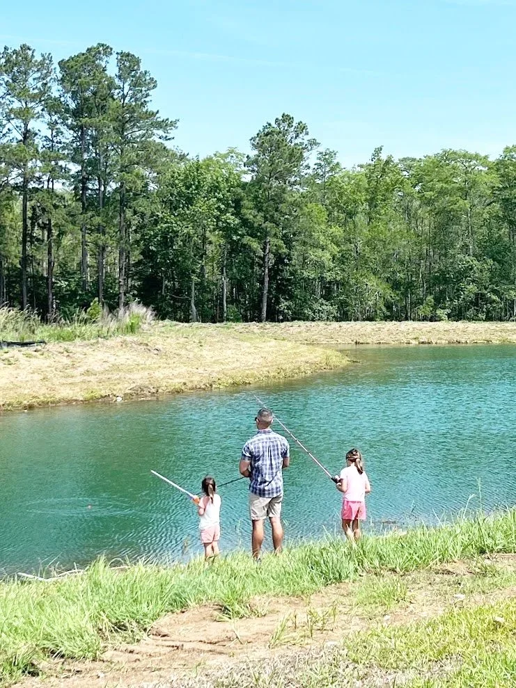 A man and two young girls fishing by a lake on a sunny day, with a forested background.