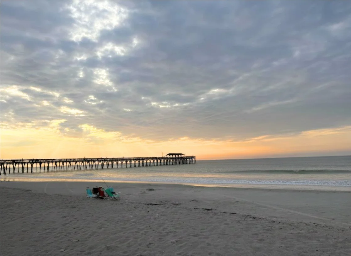 Myrtle Beach State Park Beach with a pier extending into the ocean at sunset, with chairs on the sand and cloudy sky.