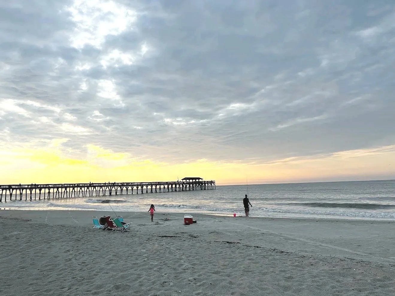 People fishing and relaxing on a sandy beach near a pier at sunset, with cloudy sky and calm ocean.