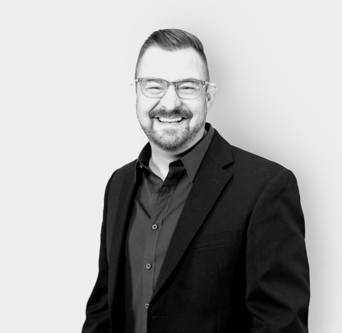 Black and white portrait of a smiling man with glasses, short hair, a beard and mustache, wearing a dark blazer and a collared shirt, against a plain light background.