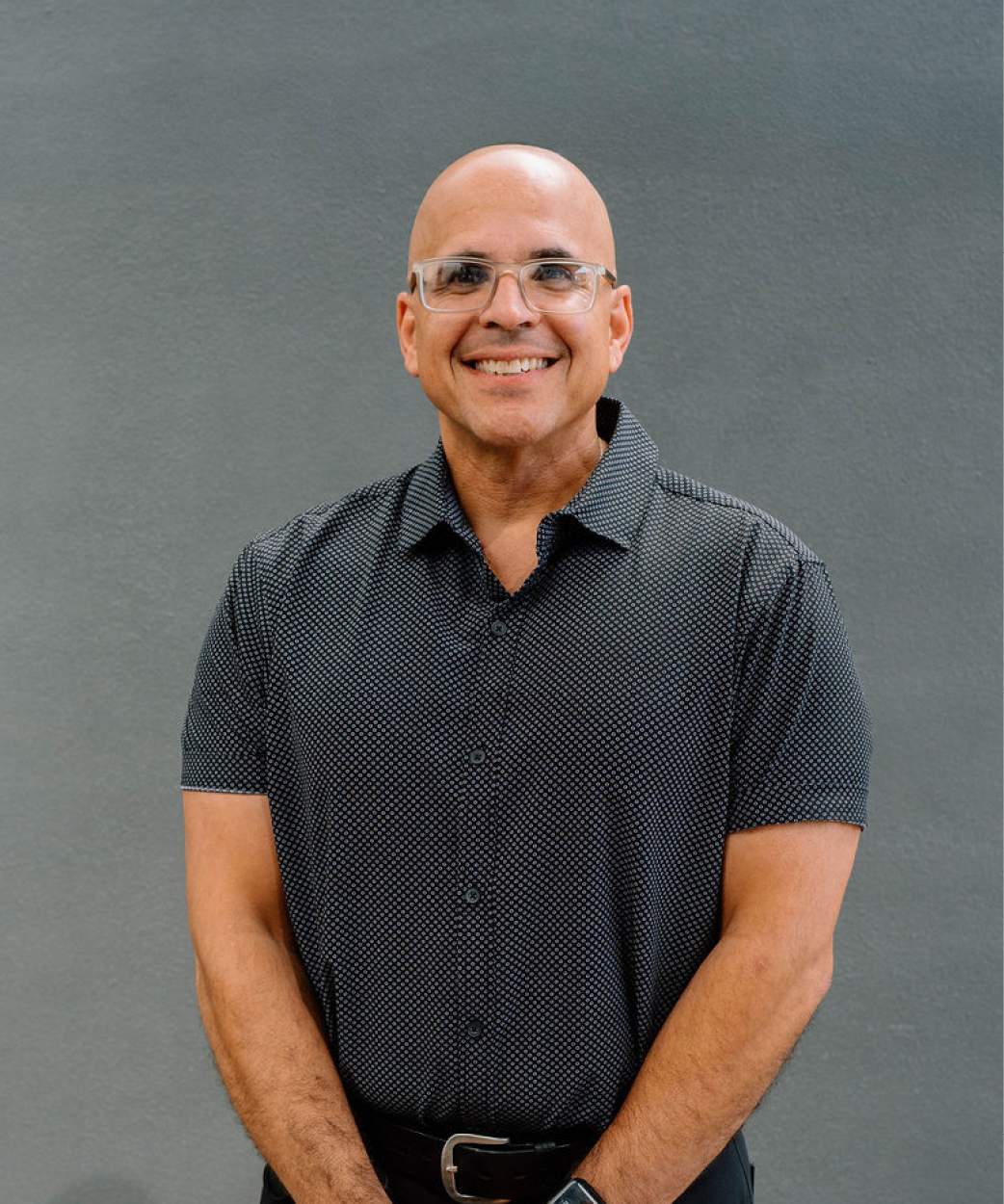 Smiling man with glasses and a shaved head, wearing a black patterned short-sleeve shirt and a watch, standing against a gray wall.