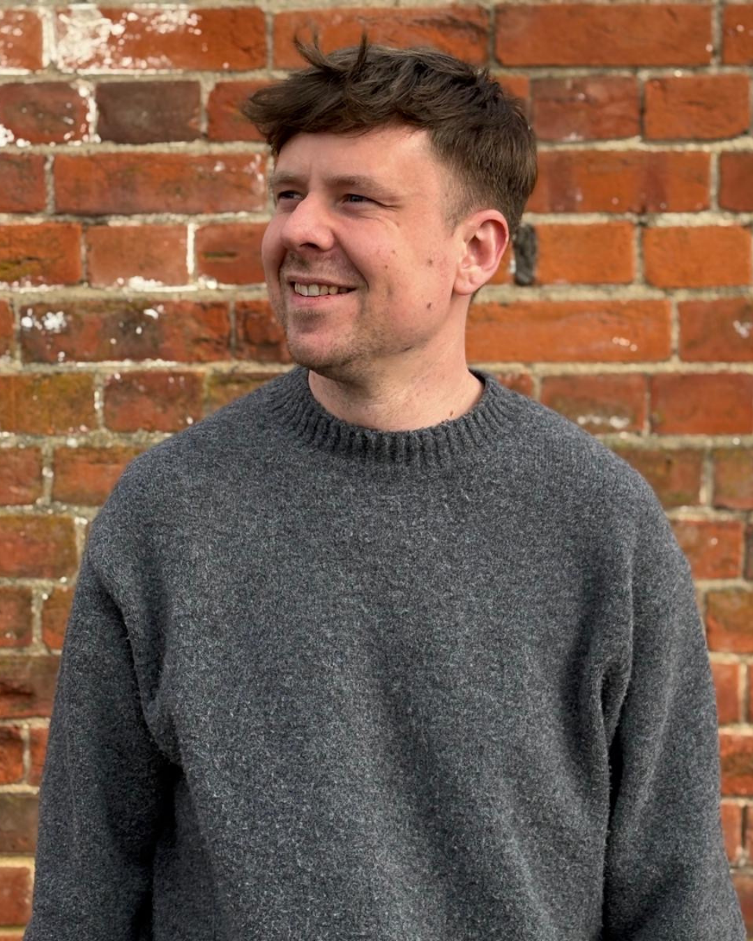 Therapist Matt Salter smiling and wearing a gray jumper standing in front of a red brick wall.