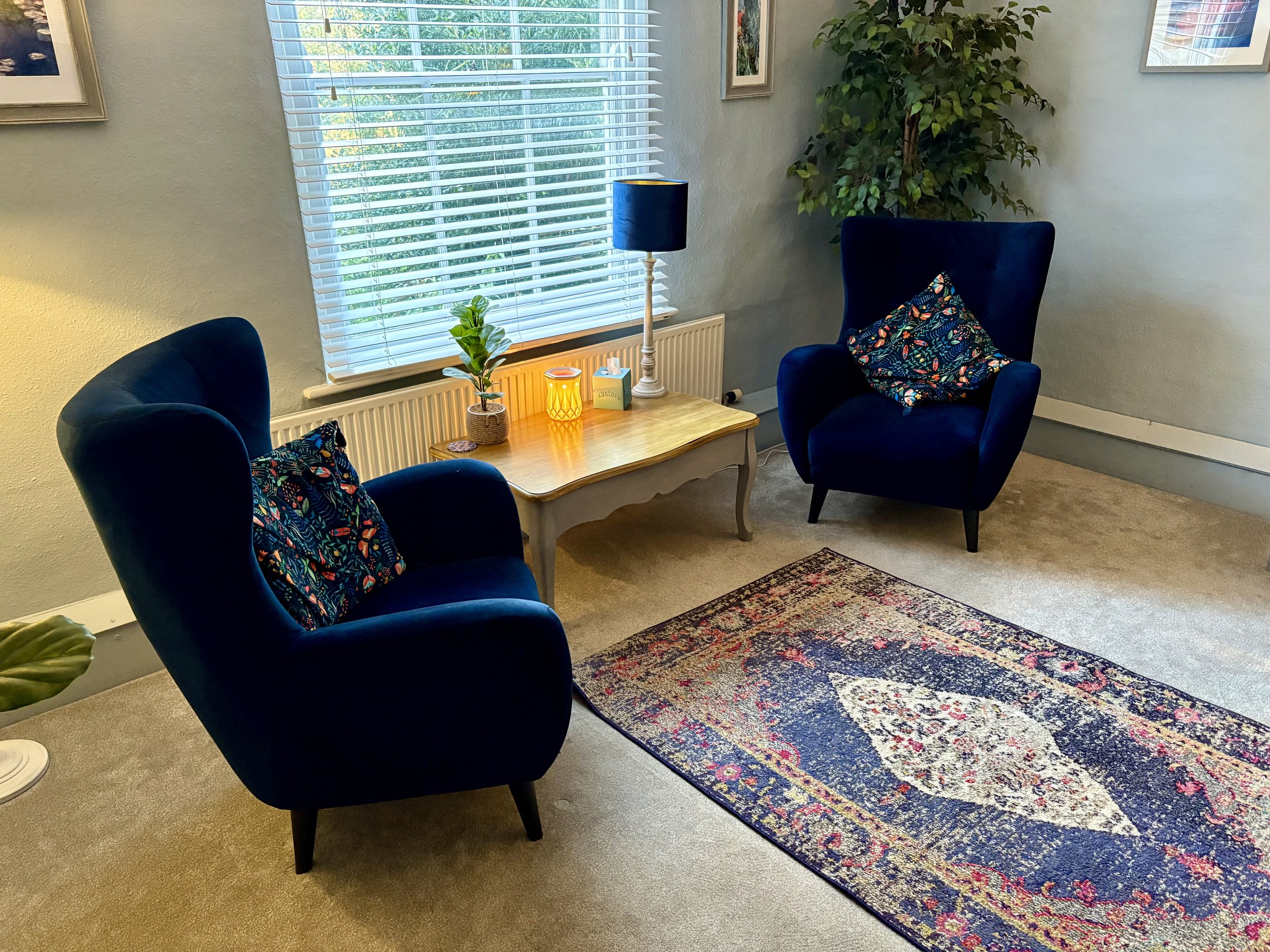 An example therapy room with two blue velvet armchairs, colorful patterned throw pillows, a wooden table, a large window with blinds, a potted plant, a table lamp, and decorative items, with a patterned rug on the beige carpeted floor.