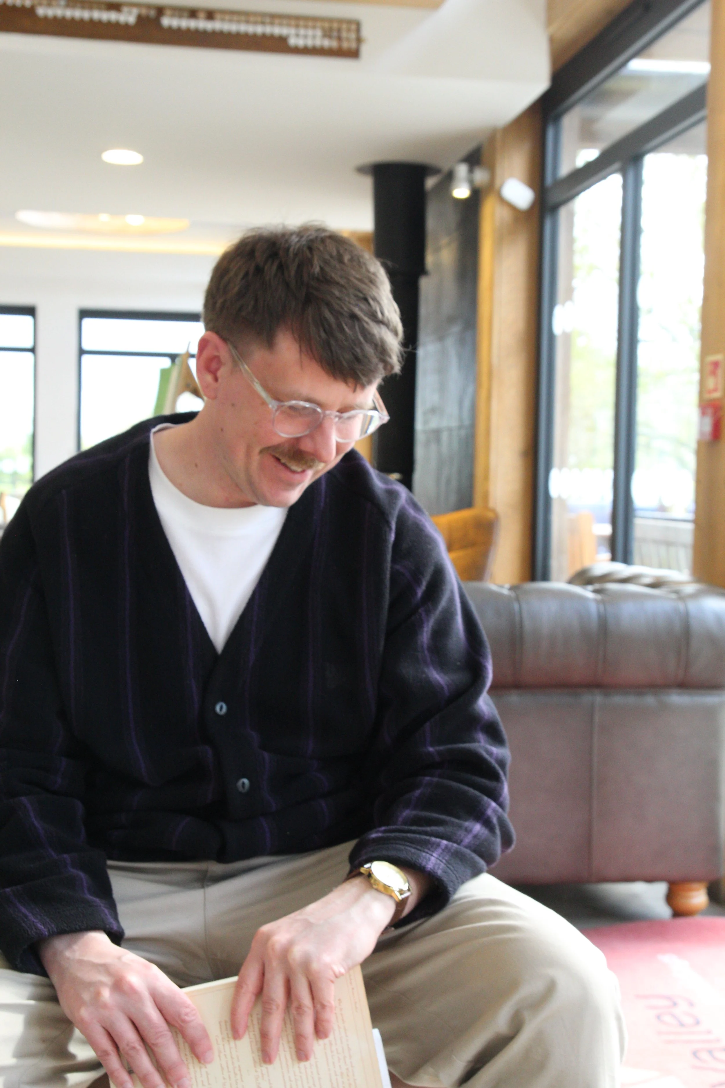 Therapist Matthew Salter with glasses and a mustache reading a book inside a cafe, with large windows and wooden walls in the background.