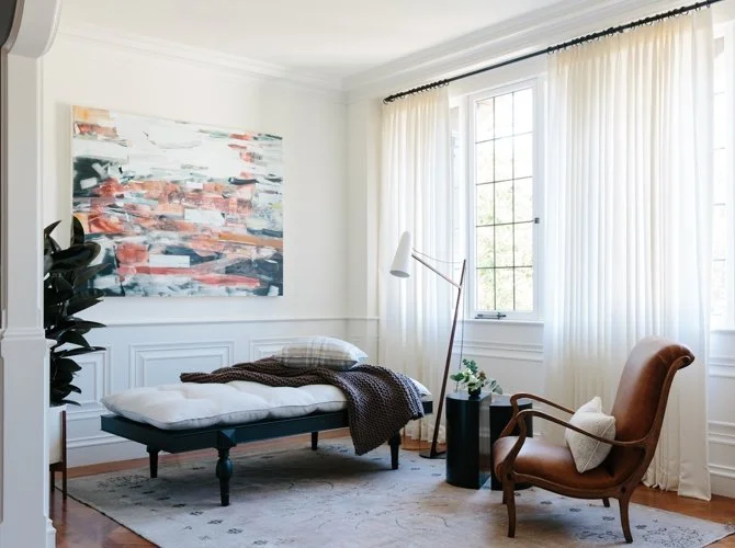 Bright bedroom with a bed, abstract painting, armchair, lamp, and window with white curtains.