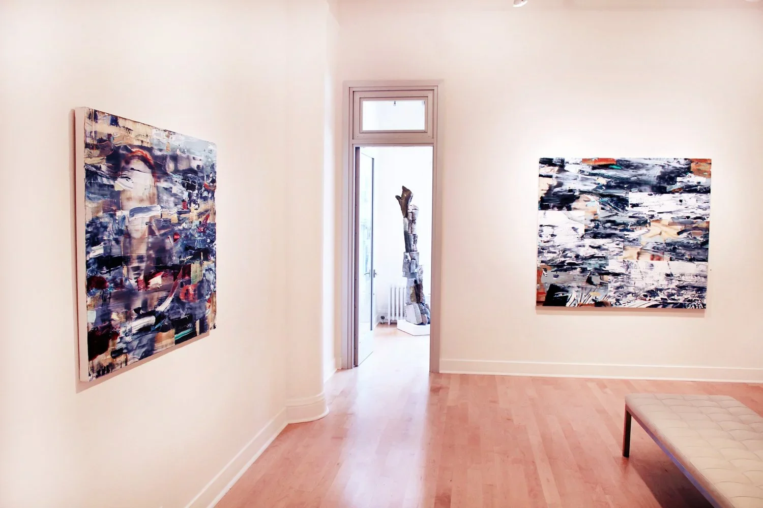 Art gallery room with two abstract paintings on white walls and a wooden floor, view through an open door revealing a sculpture in another room.