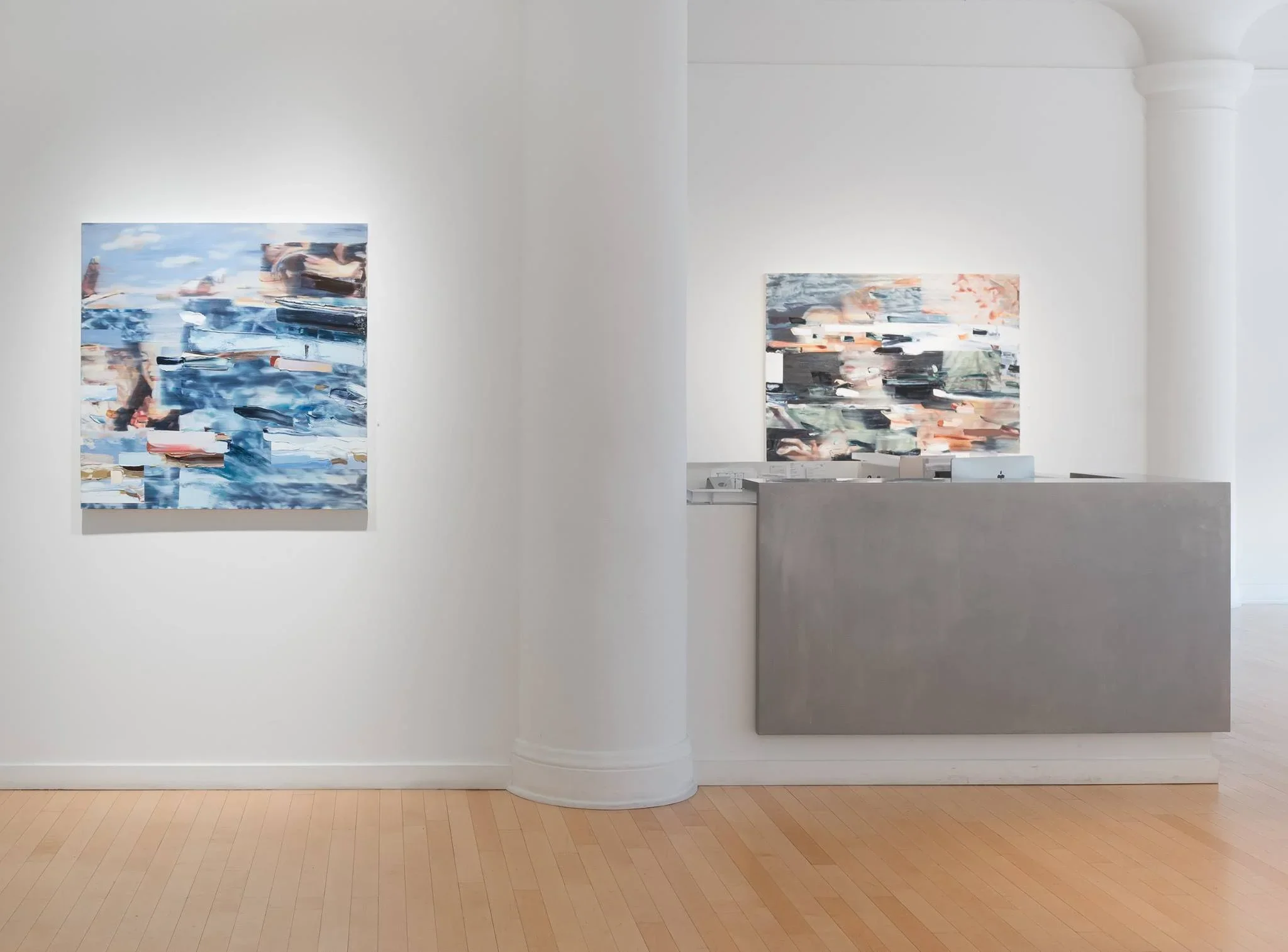 Abstract paintings displayed on white gallery walls with a large column in the corner, hardwood floor, and minimalist setting.