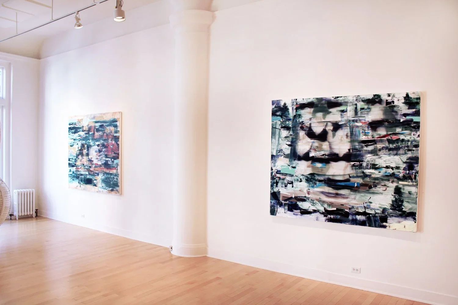 An art gallery with white walls and wooden floors displaying abstract paintings, one of which features a black and white portrait with vibrant brushstrokes.