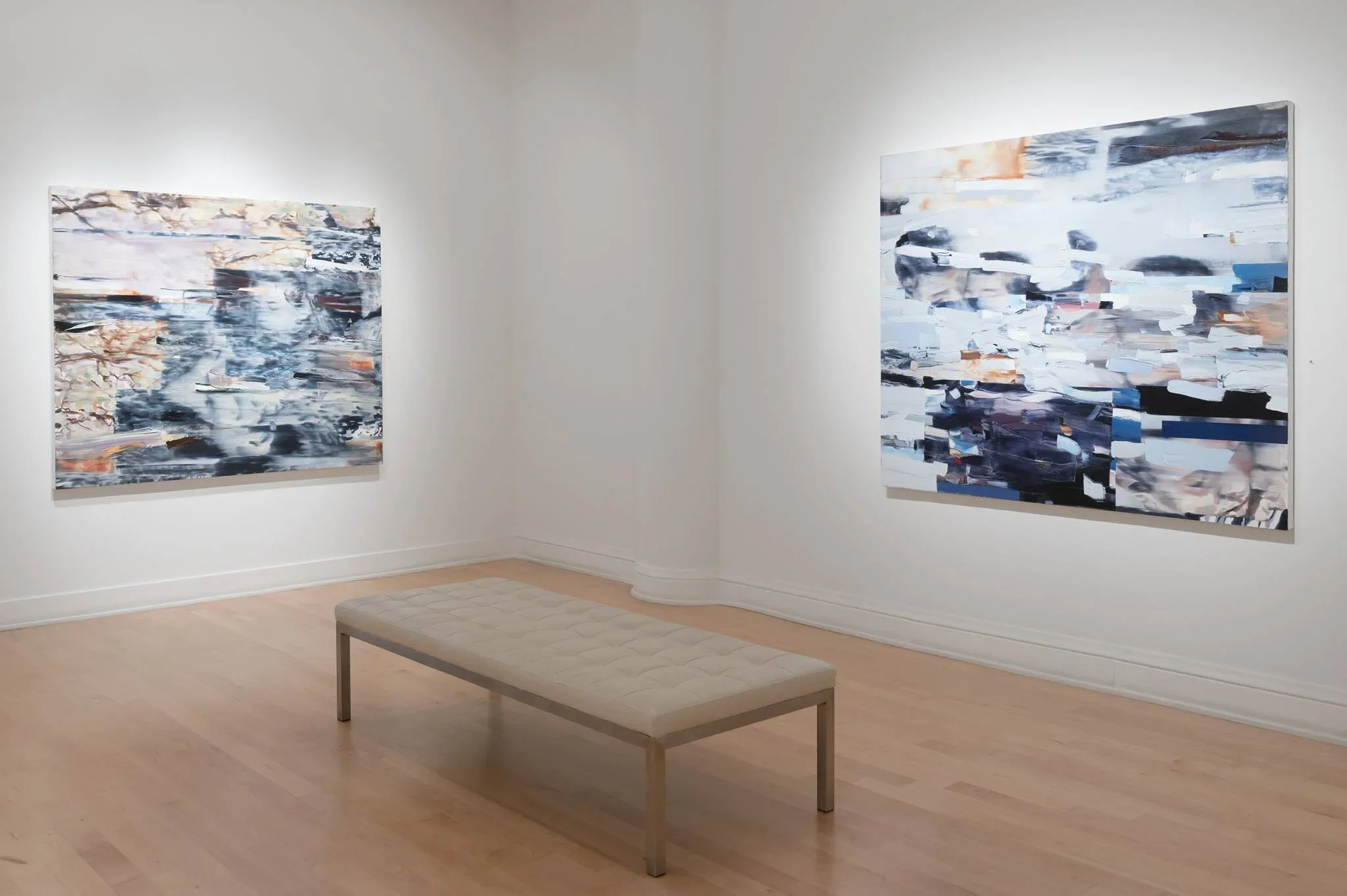 Two abstract paintings displayed on a white wall in an art gallery. A beige bench is in the foreground, and the gallery has light wooden flooring.