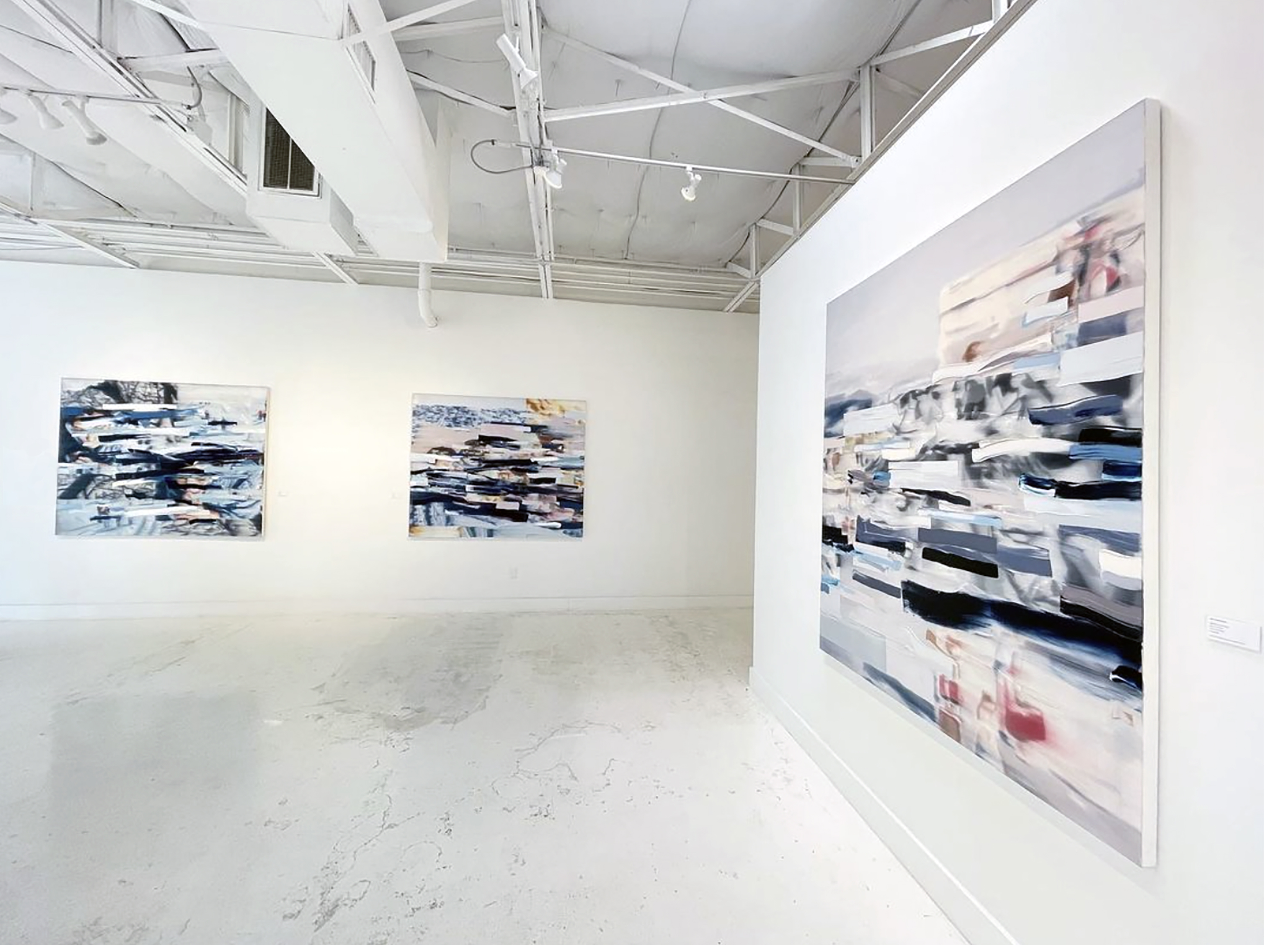 Art gallery with abstract paintings on white walls and a high ceiling with exposed ductwork and lighting.