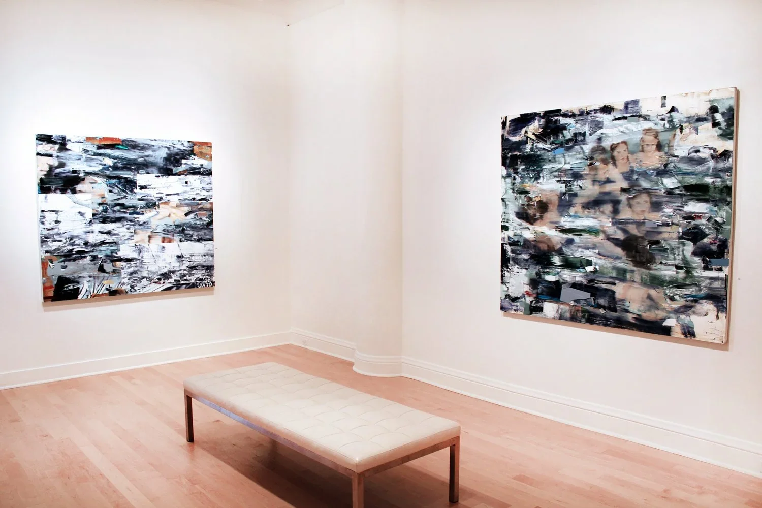 An art gallery displaying two abstract paintings on white walls and a cream-colored bench on a light wooden floor.