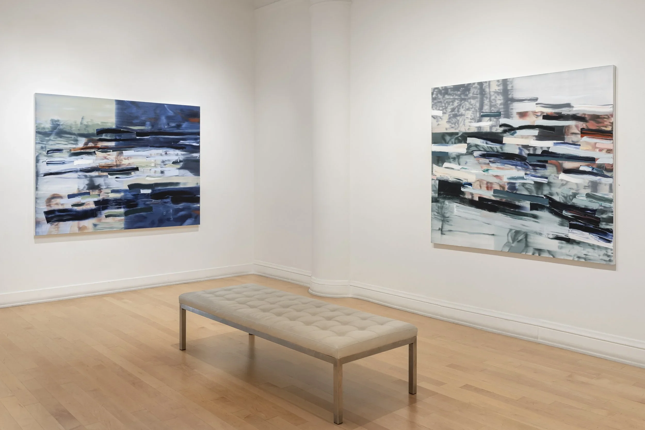 Two abstract paintings hang on white gallery walls, featuring blurred horizontal brushstrokes in shades of blue, gray, black, and hints of rust. A light gray upholstered bench sits in front of the paintings on a wooden floor.