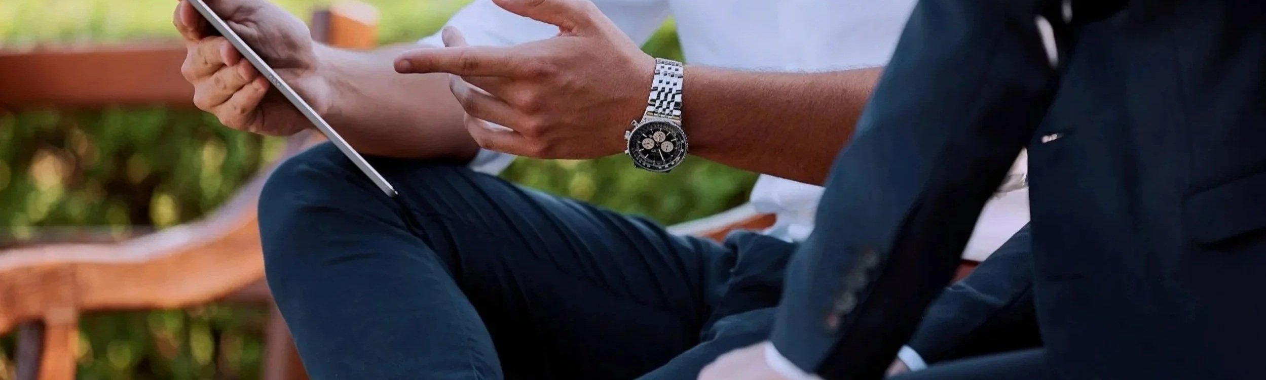 Close-up of a man using a tablet, wearing a silver watch and dark clothing, sitting outdoors.