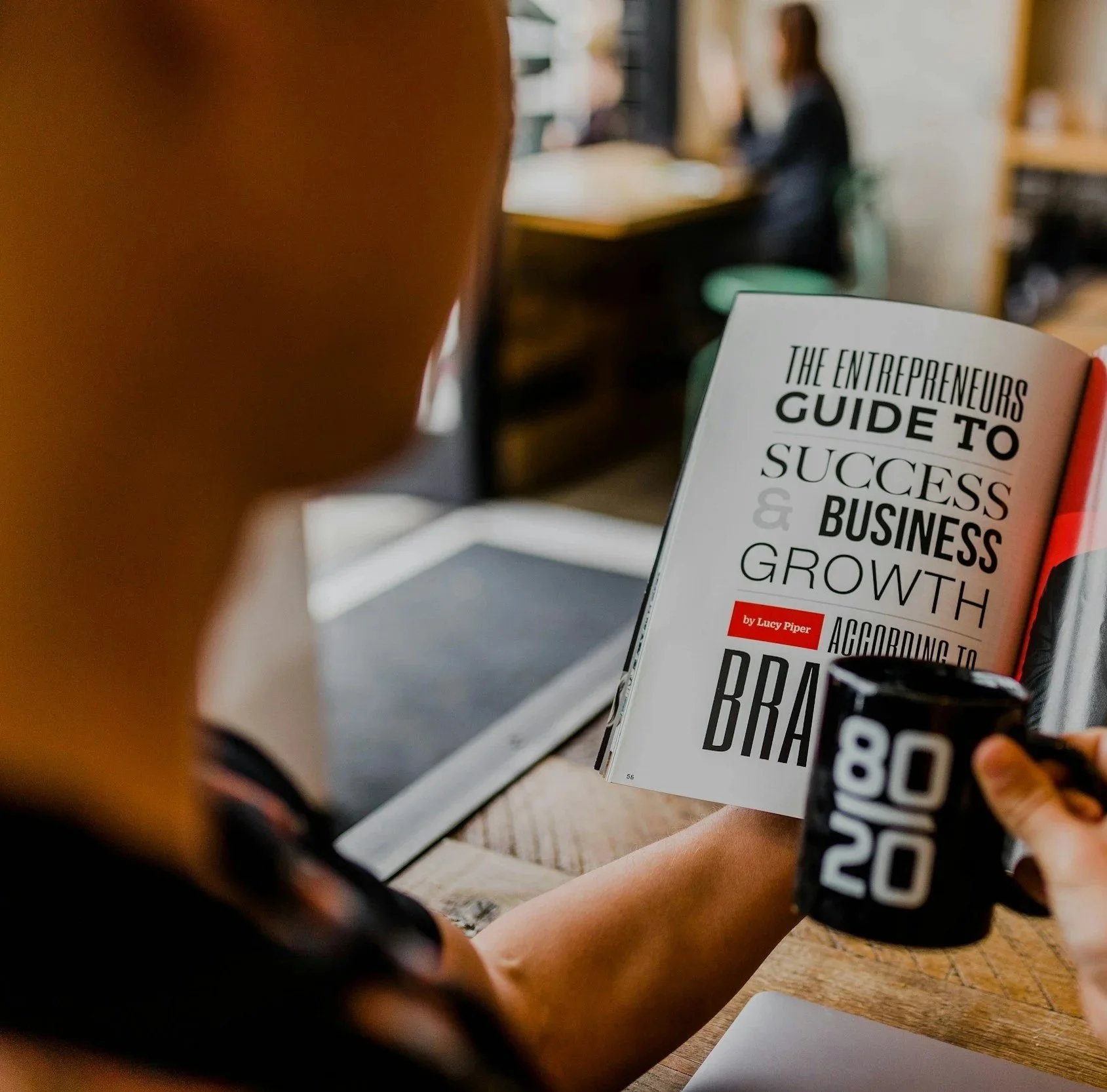 Person sitting at a table reading a book titled 'The Entrepreneurs Guide to Success & Business Growth' while holding a mug with the text '80 90 100' on it, in a cozy indoor setting.