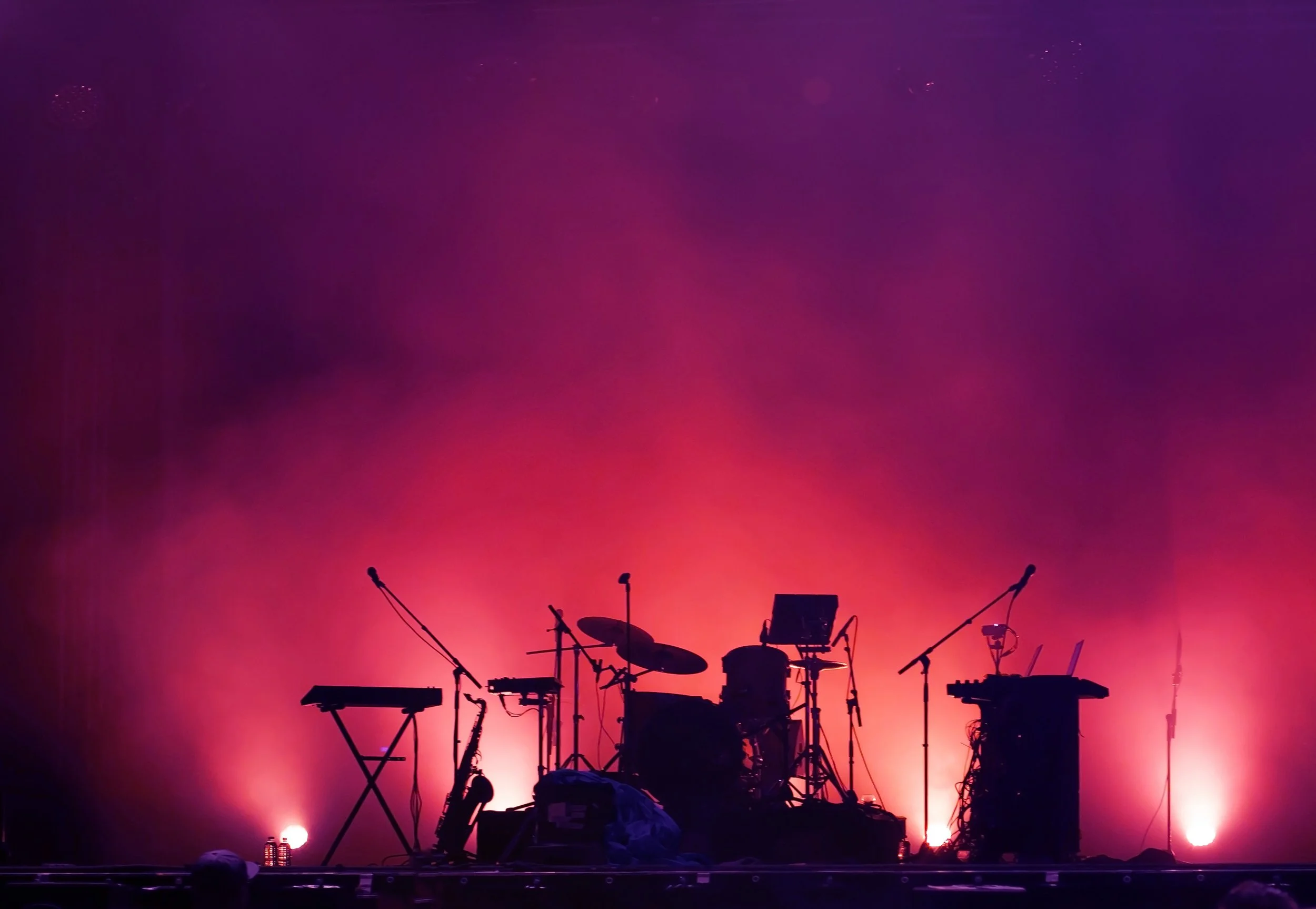 Silhouette of musical instruments, including drums, keyboards, and microphones, set against a vibrant purple and pink gradient stage lighting.