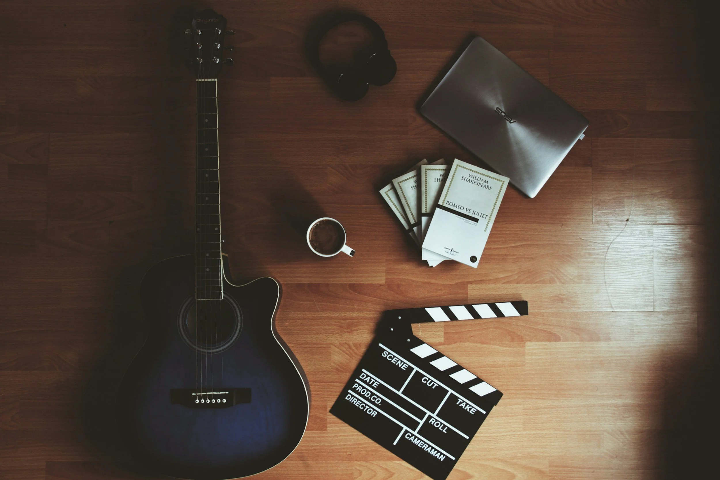 Flat lay of a black acoustic guitar, a pair of black headphones, a closed silver laptop, a stack of three William Shakespeare's "Romeo and Juliet" books, a paper coffee cup, and a black and white film slate on a wooden floor.