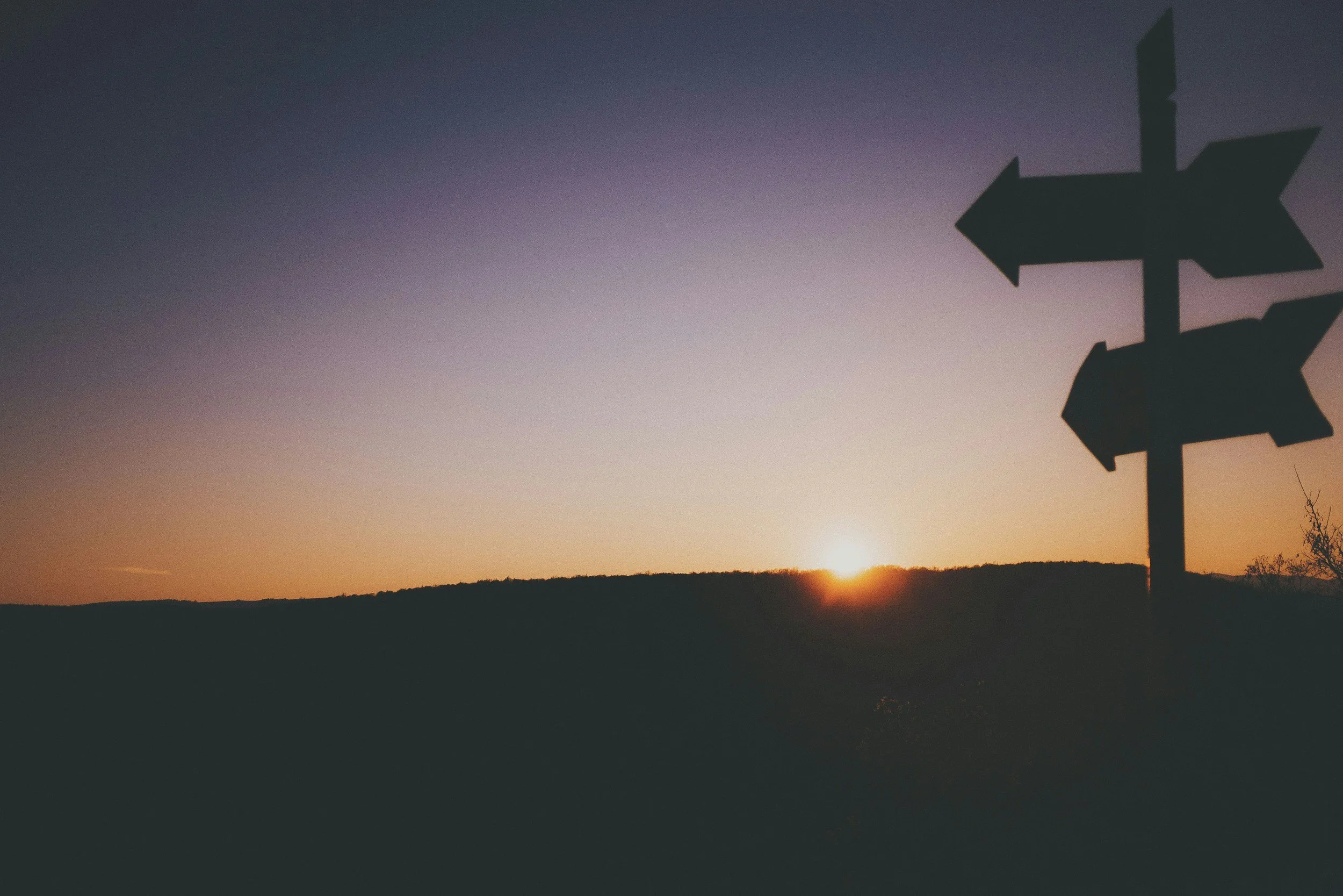 Silhouette of a signpost with multiple arrows against a colorful sunset sky with a low horizon and a few trees in the distance.
