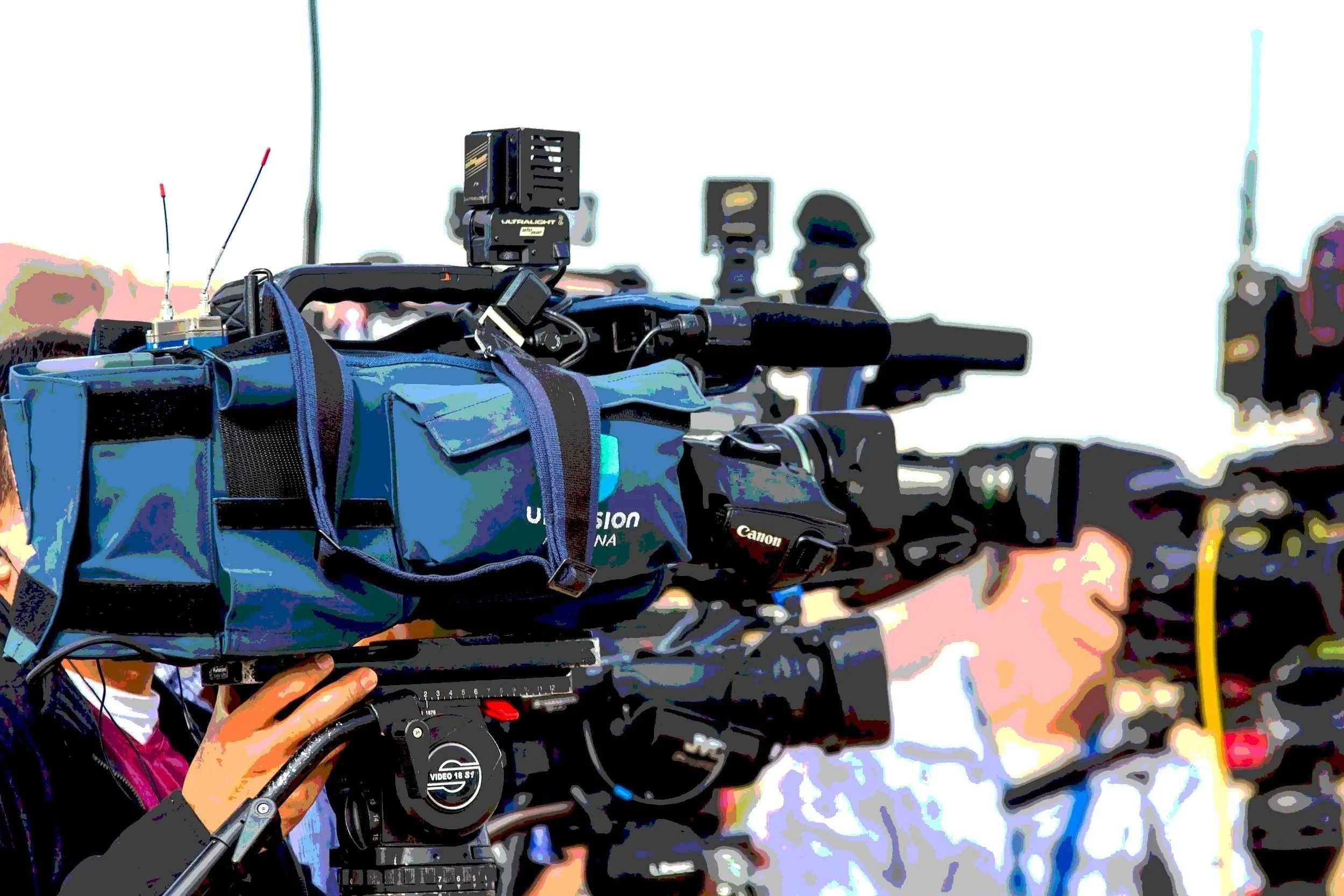 Close-up of a person operating a professional video camera mounted on a tripod, with additional equipment and wires attached, outdoors during daytime.