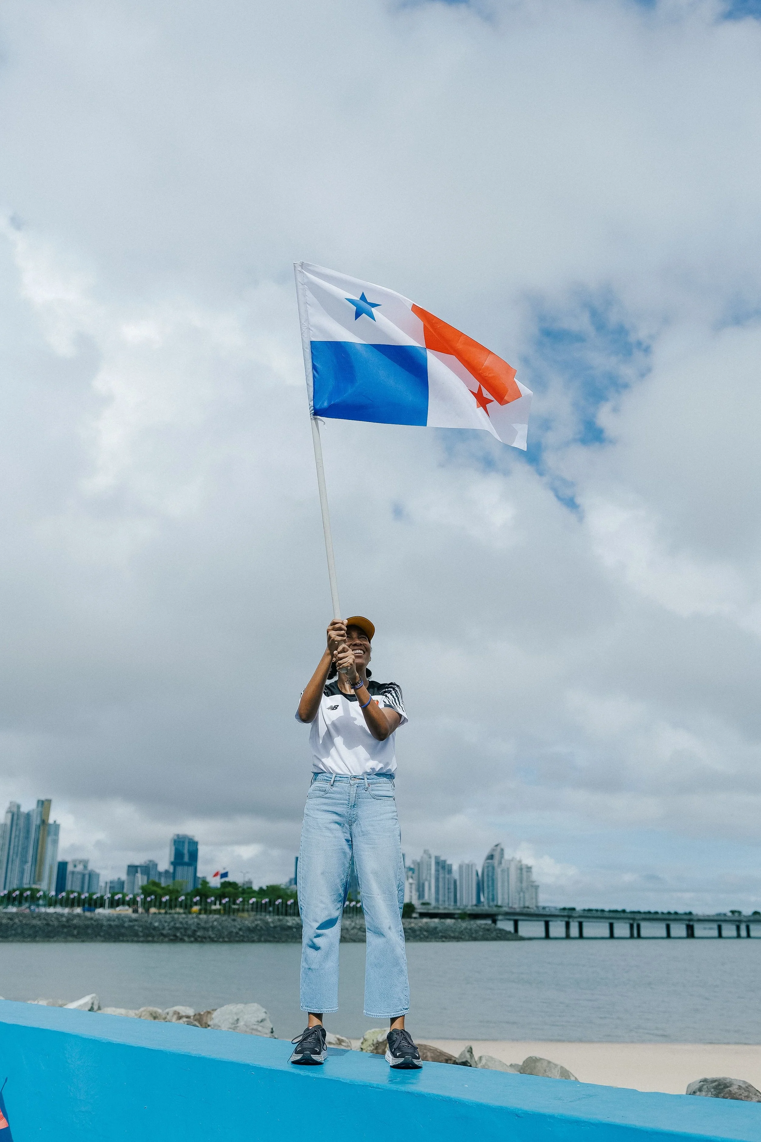 Person standing on a ledge by the water, holding a flag with blue, red, and white colors. City skyline in the background under a cloudy sky.