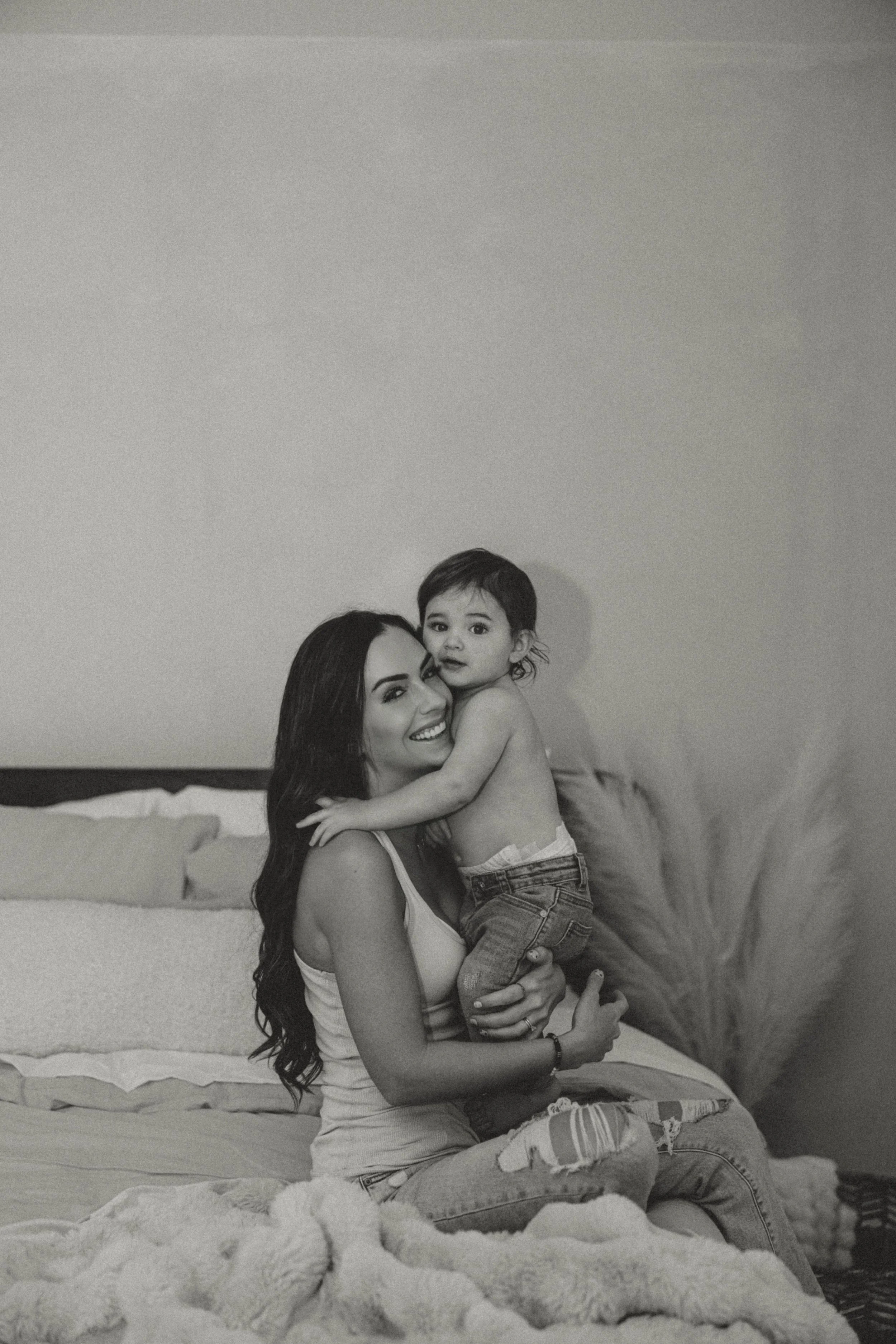 A woman with long dark hair and a child with short dark hair embrace on a bed in a bedroom. The woman is smiling and holding the child, who is looking at the camera. The room has a plain wall and a stack of blankets or clothing on the bed.