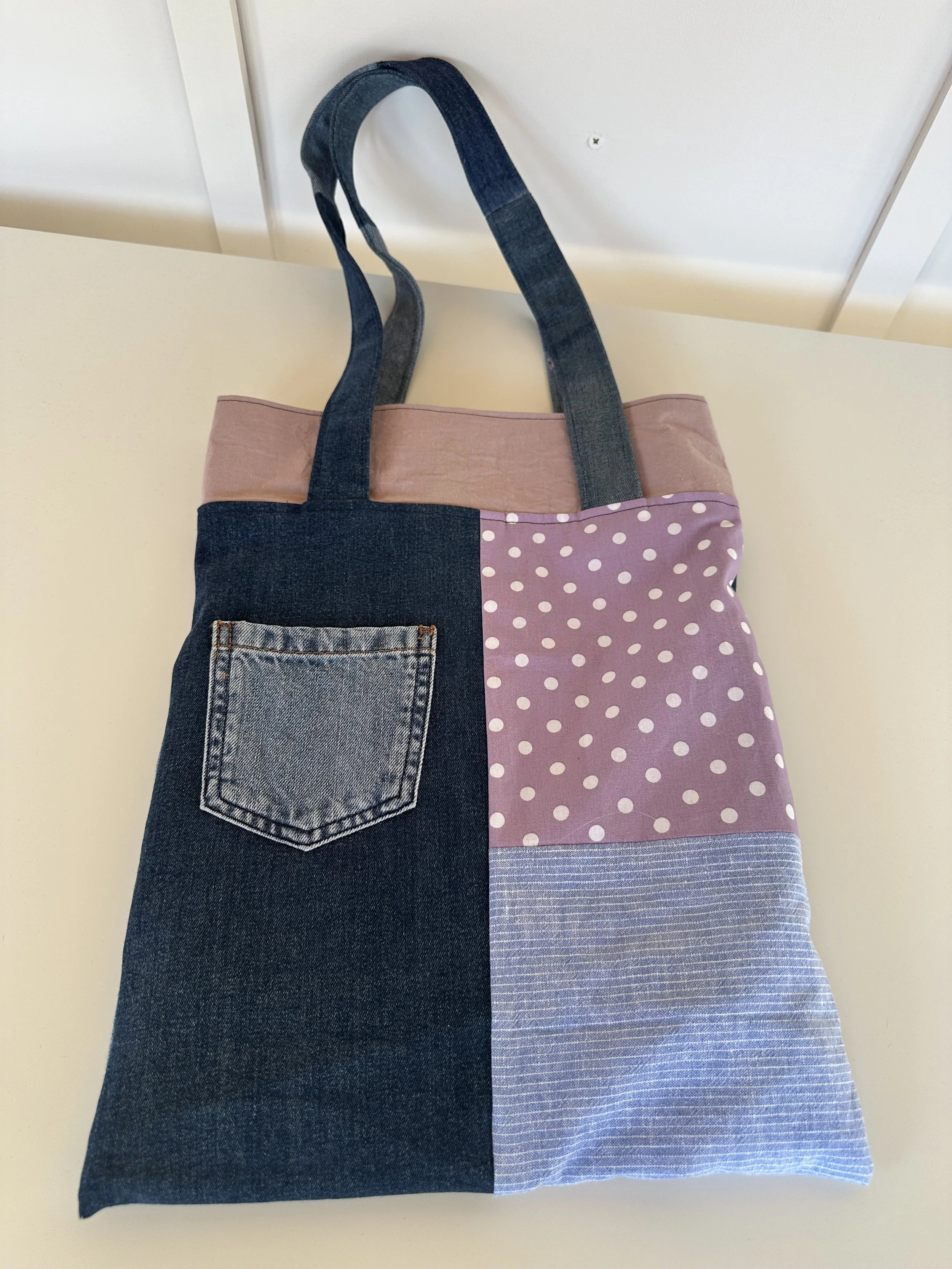 A tote bag made of denim and fabric, with one side featuring a denim pocket and the other side a pink polka dot and striped pattern, with dark denim straps.