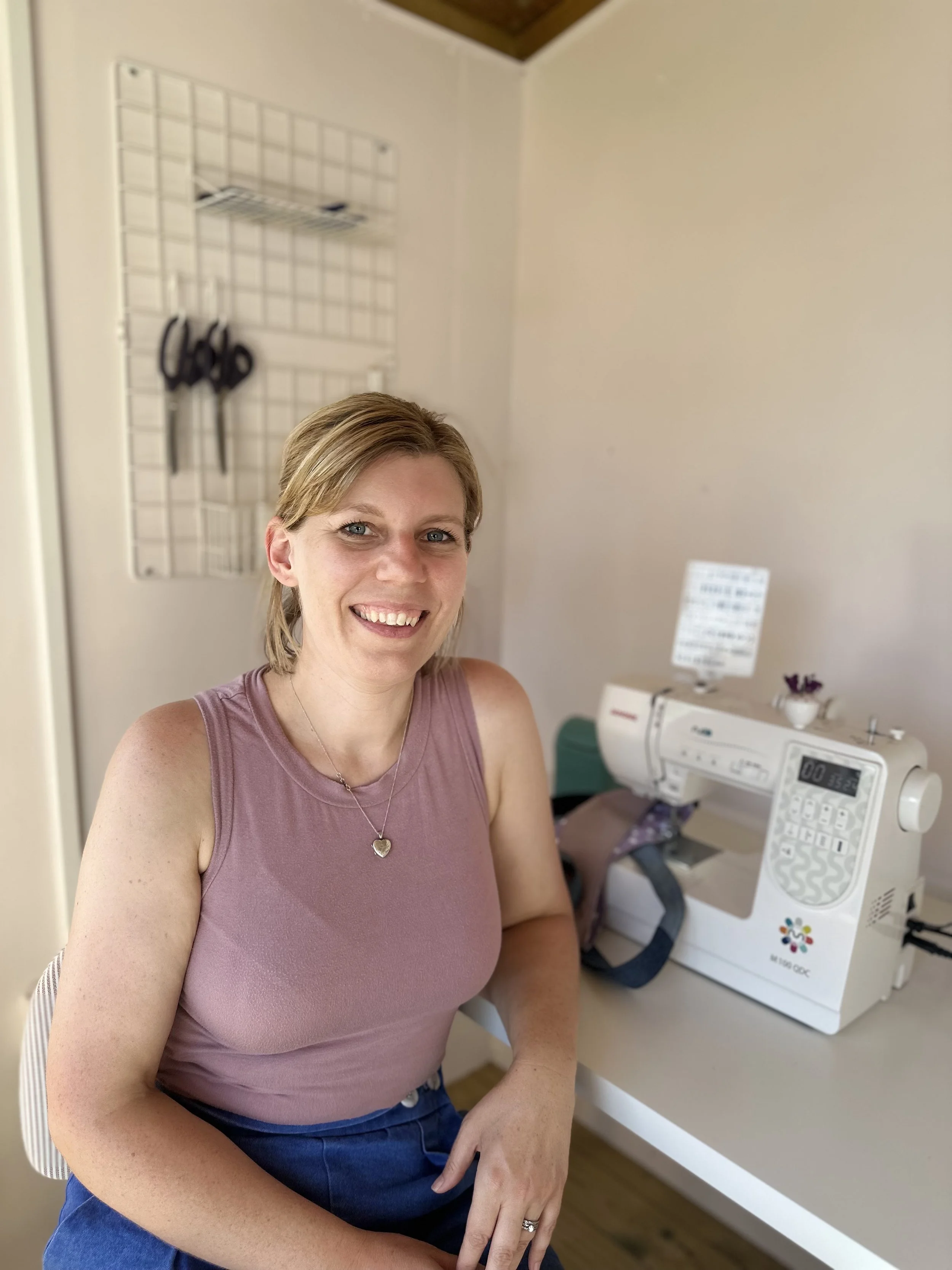 A woman with blond hair, wearing a pink sleeveless top, smiling while sitting at a sewing machine in a room with beige walls.