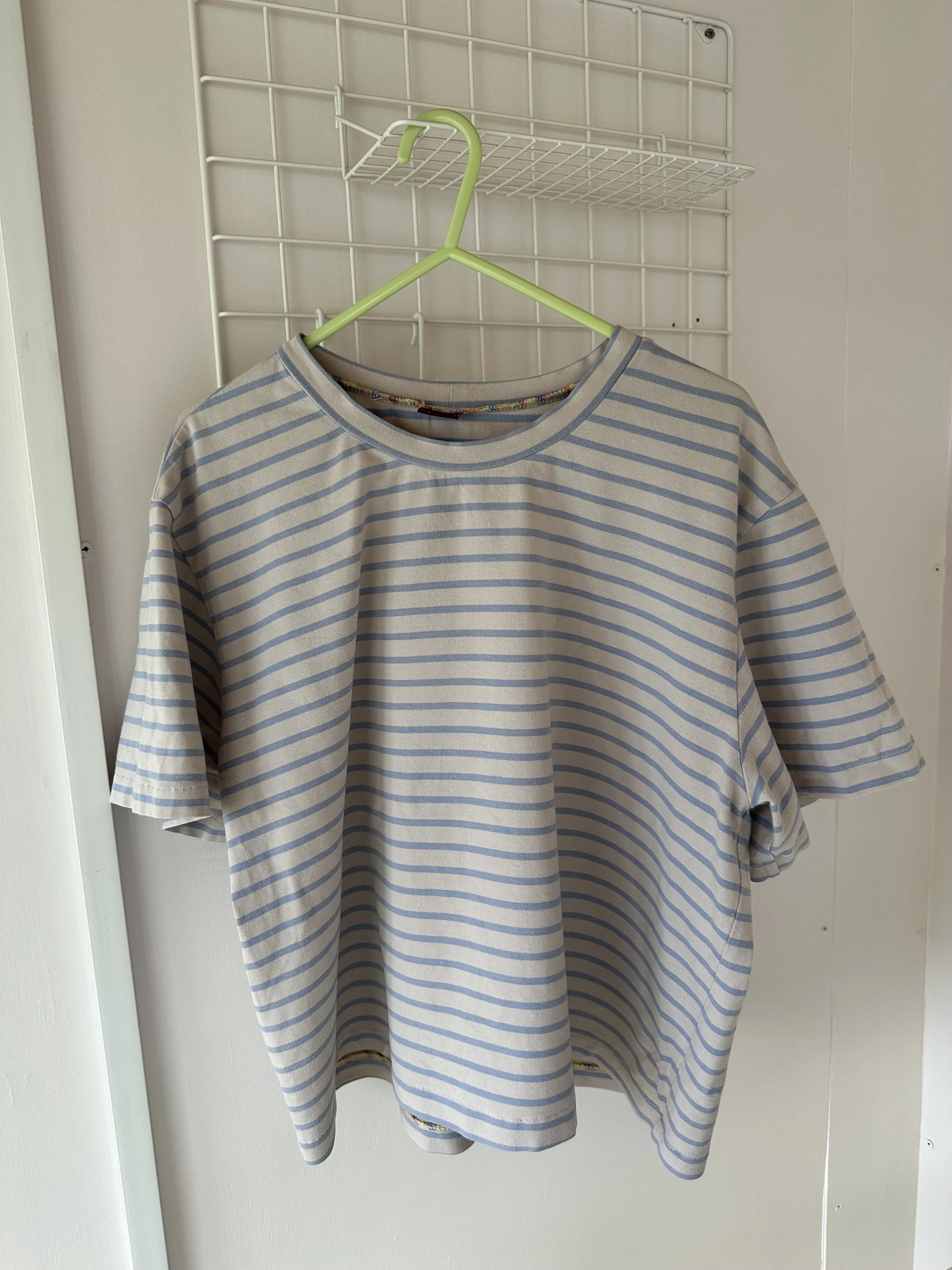 A beige and blue striped long-sleeve shirt hanging on a green hanger on a white wire grid wall.
