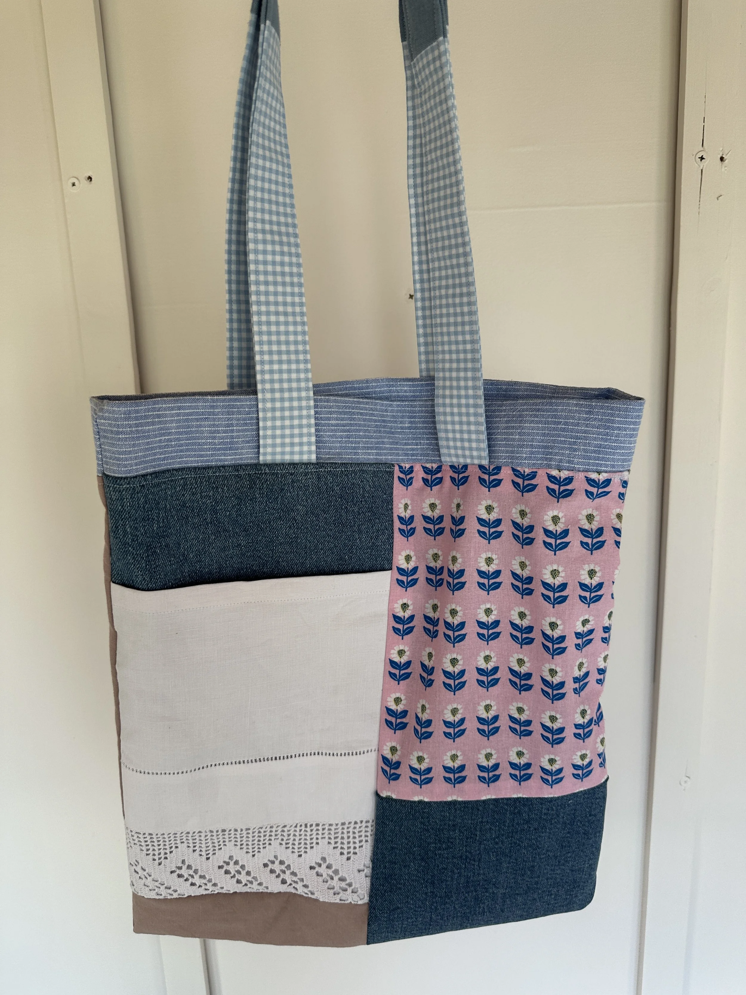 A tote bag with a patchwork design including denim, floral, and lace patterns, hanging against a white wall.