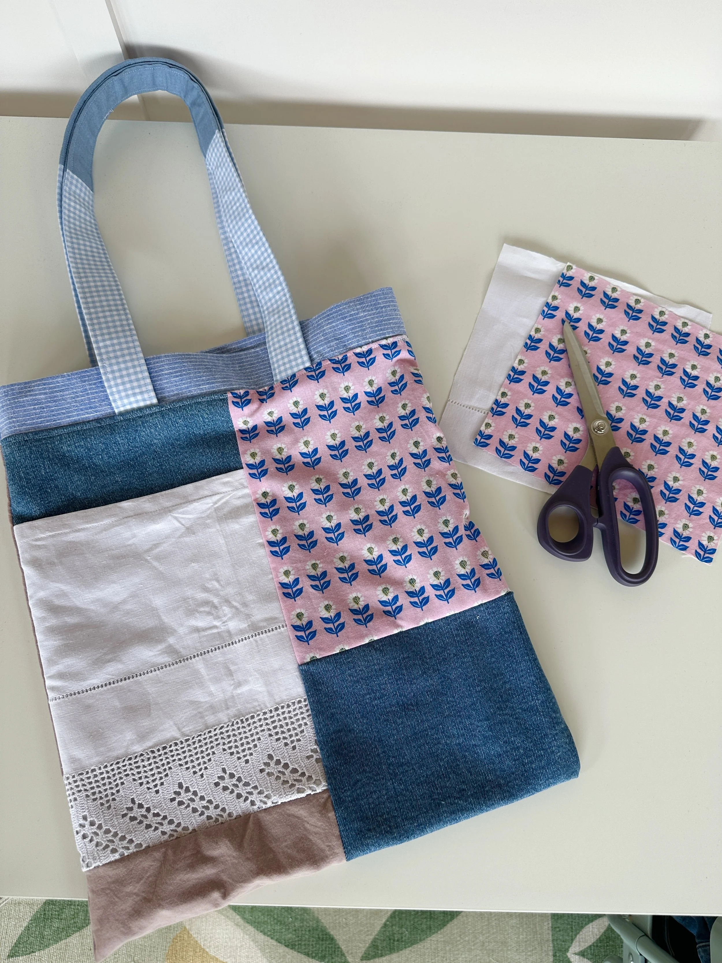 A quilted fabric bag with blue, pink, white, and denim patterns, and a pair of scissors placed on matching fabric, all on a white surface.