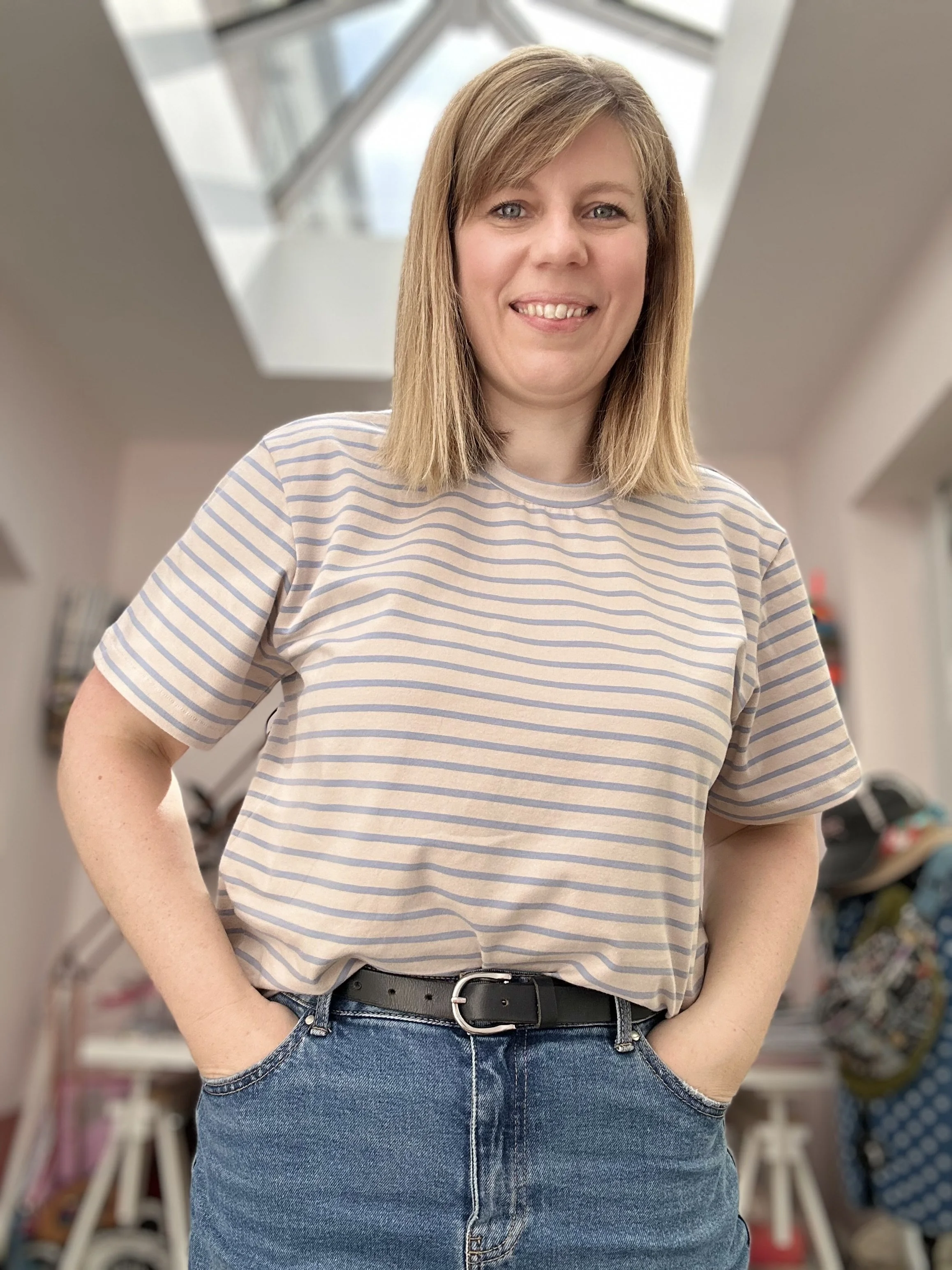 A woman with shoulder-length blonde hair, wearing a striped t-shirt and blue jeans, standing indoors in front of a blurred background.