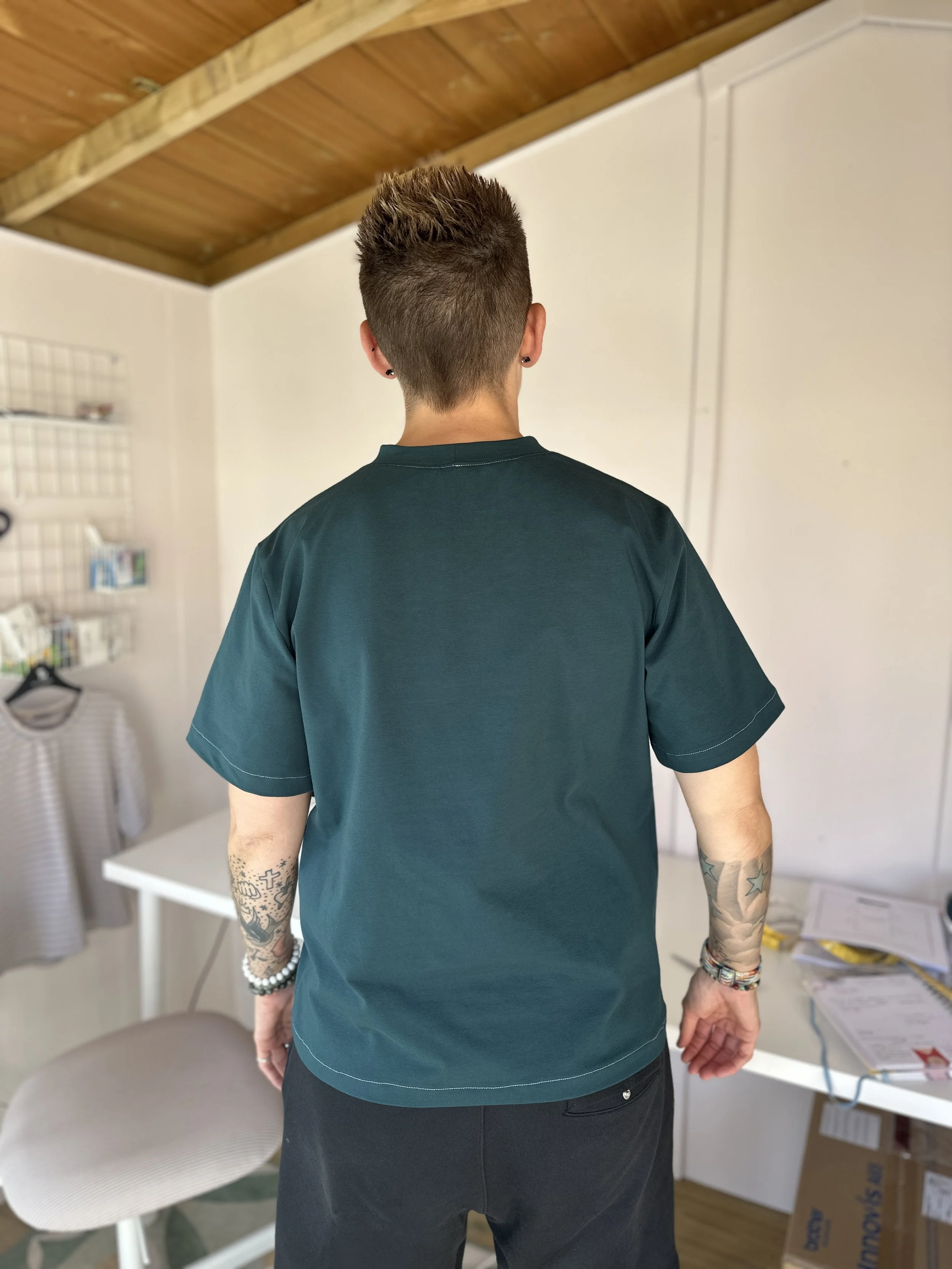 Back view of a person with short, styled hair, wearing a dark green shirt and black pants, tattoos on both arms, standing indoors near a white table and shelf, with a wooden ceiling overhead.