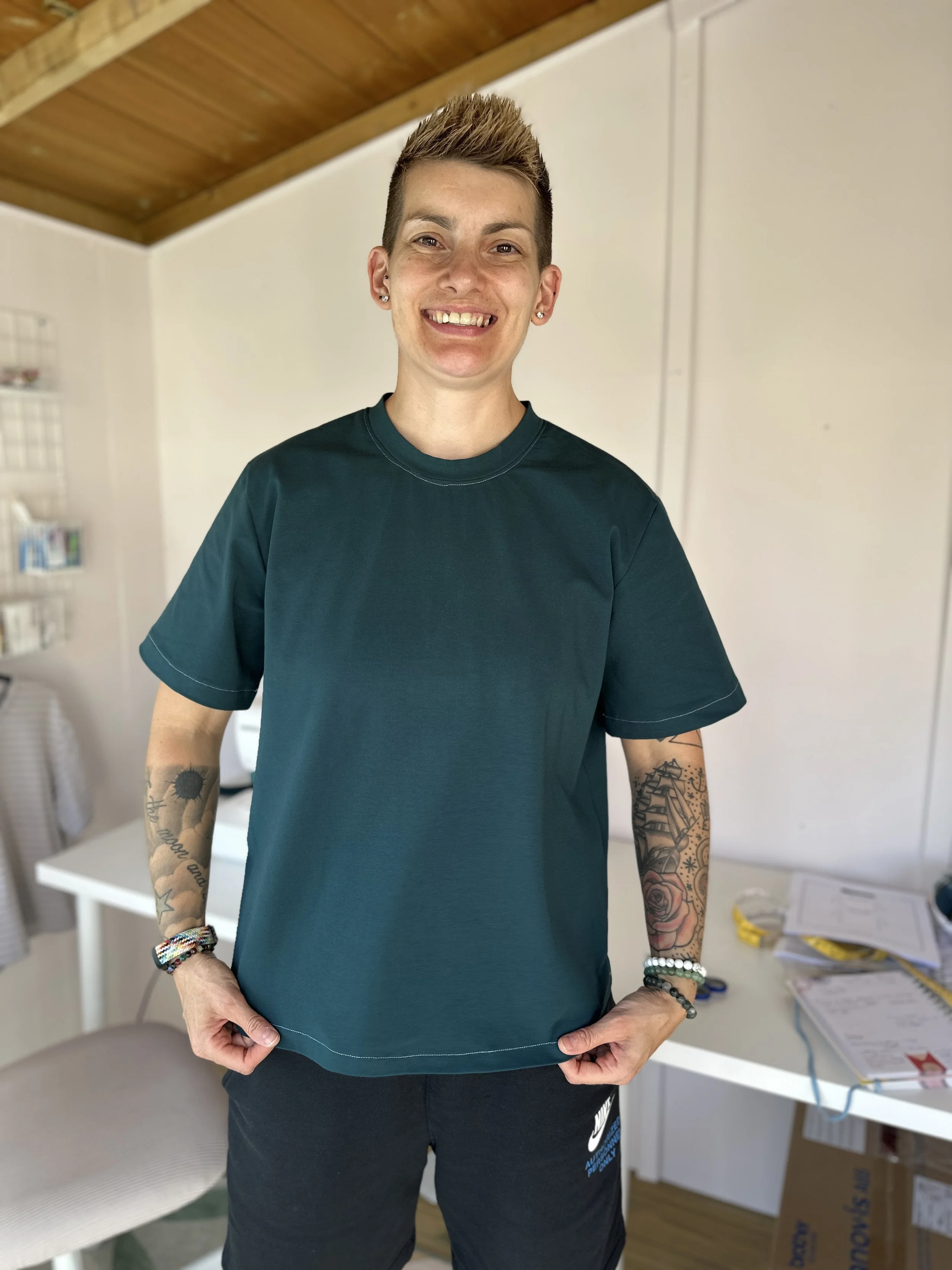 A person with short light brown hair and tattoos on both arms stands indoors, smiling at the camera. They are wearing a green t-shirt and black Nike shorts, with several bracelets on their wrists.