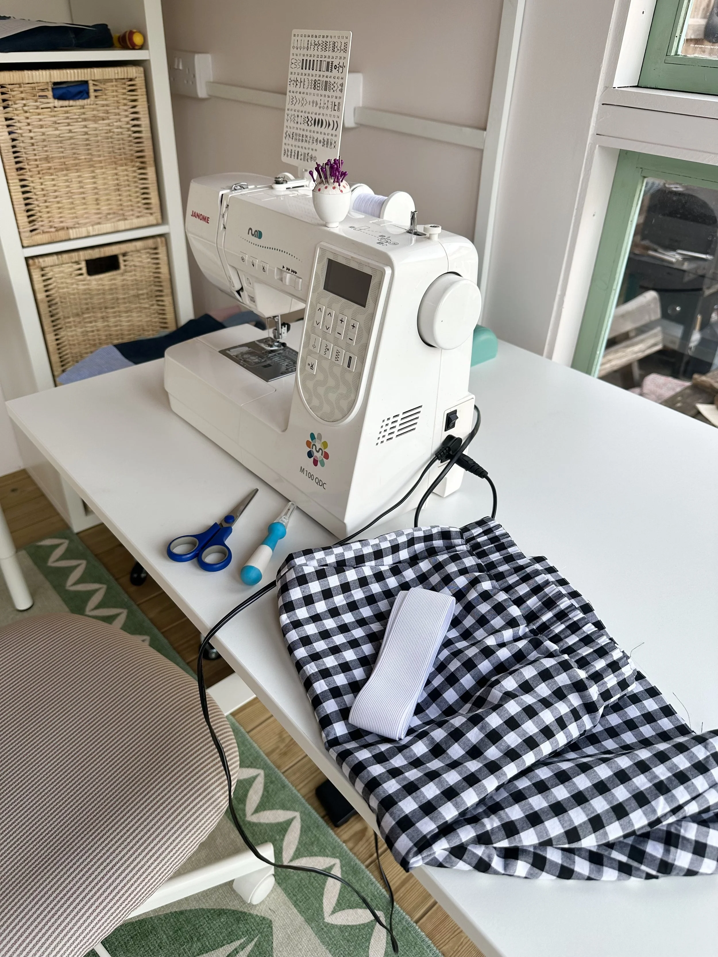 A sewing machine on a white table with fabric, scissors, and other sewing tools, near a window and a chair with a patterned cushion.