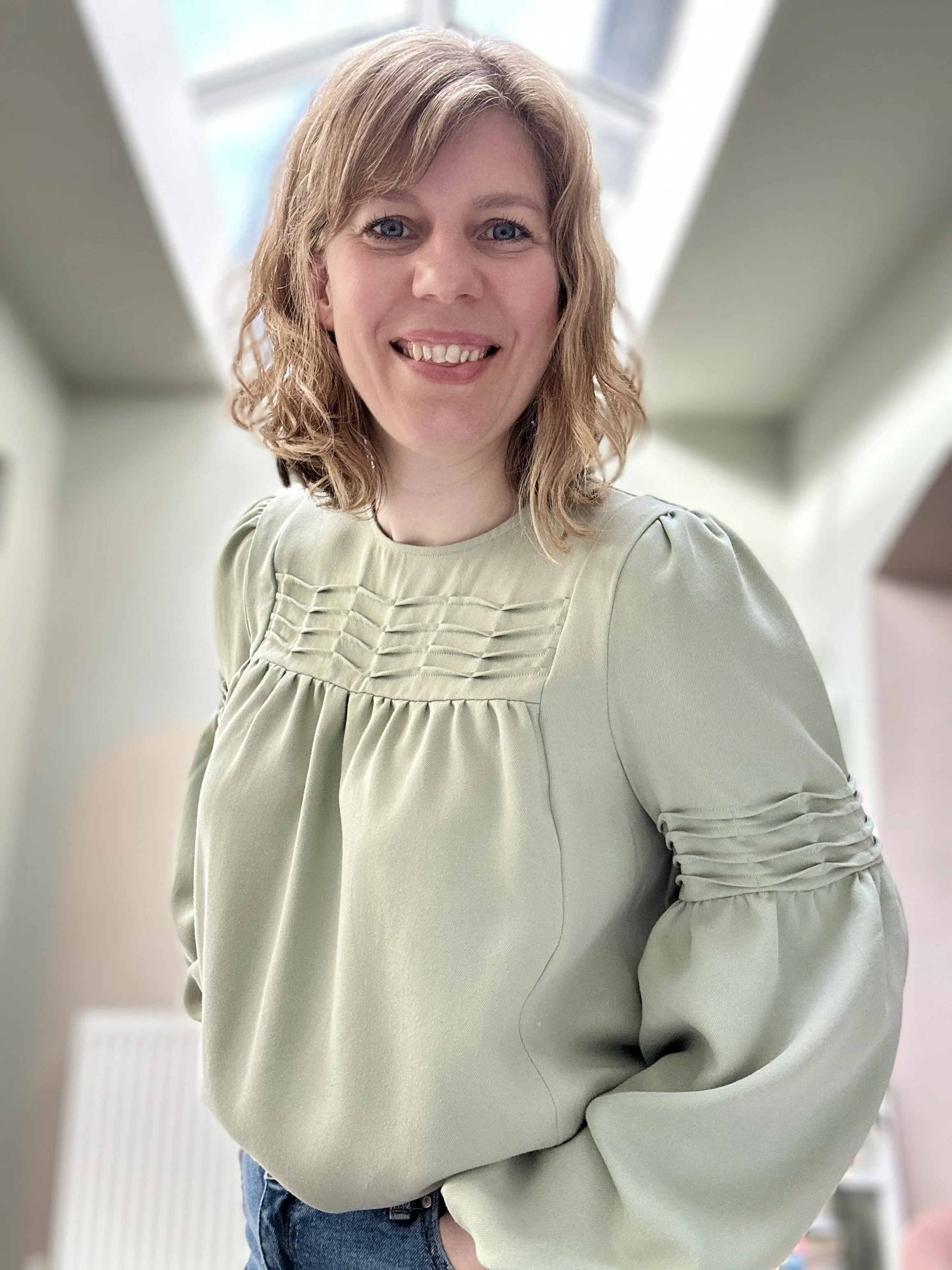 A woman with shoulder-length wavy blonde hair, blue eyes, and a wide smile, wearing a light green blouse with gathered details on the bodice and sleeves, standing in a bright indoor space with a window in the background.