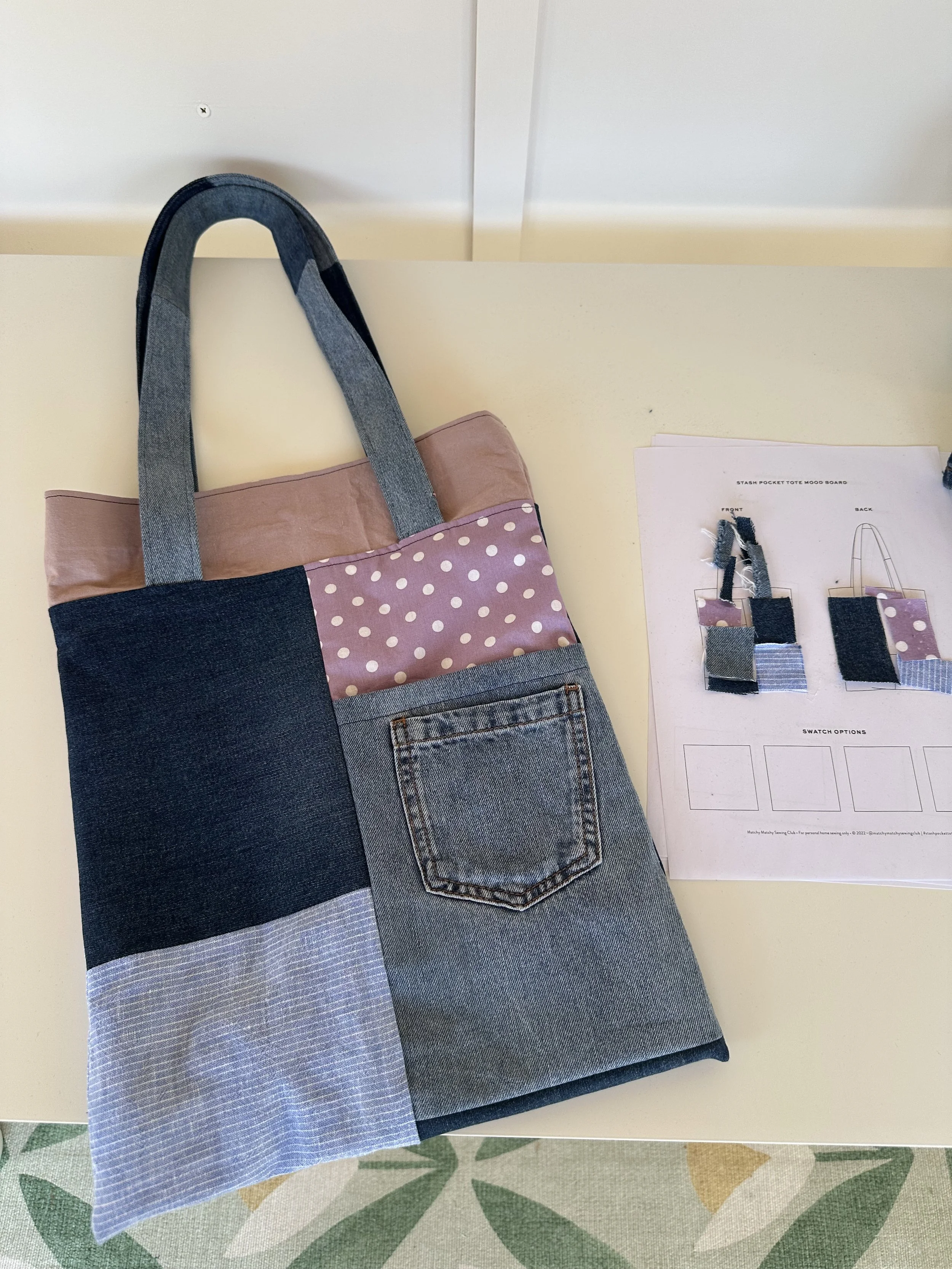 A tote bag made from denim, pink polka-dot fabric, and light blue striped fabric, placed on a table next to a printed sewing pattern.