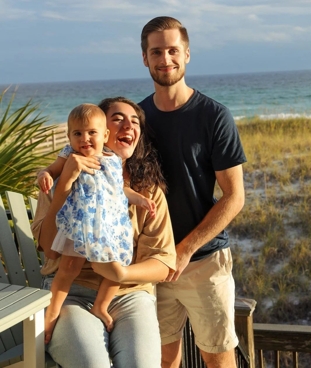 Three people on a beach, a woman holding a young girl, a man standing beside them, all smiling, with the ocean and grassy dunes in the background.