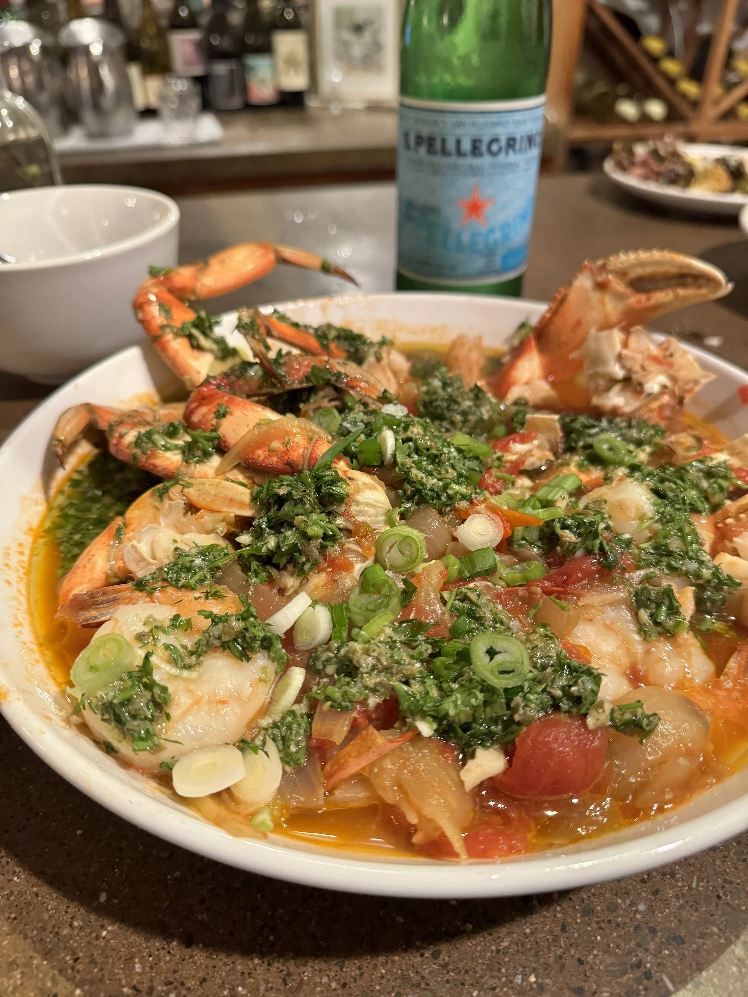 A bowl of seafood boil with shrimp, crab, sausage, corn, and vegetables garnished with green onions, on a dark countertop. In the background, a green bottled beverage and other dinnerware are visible.