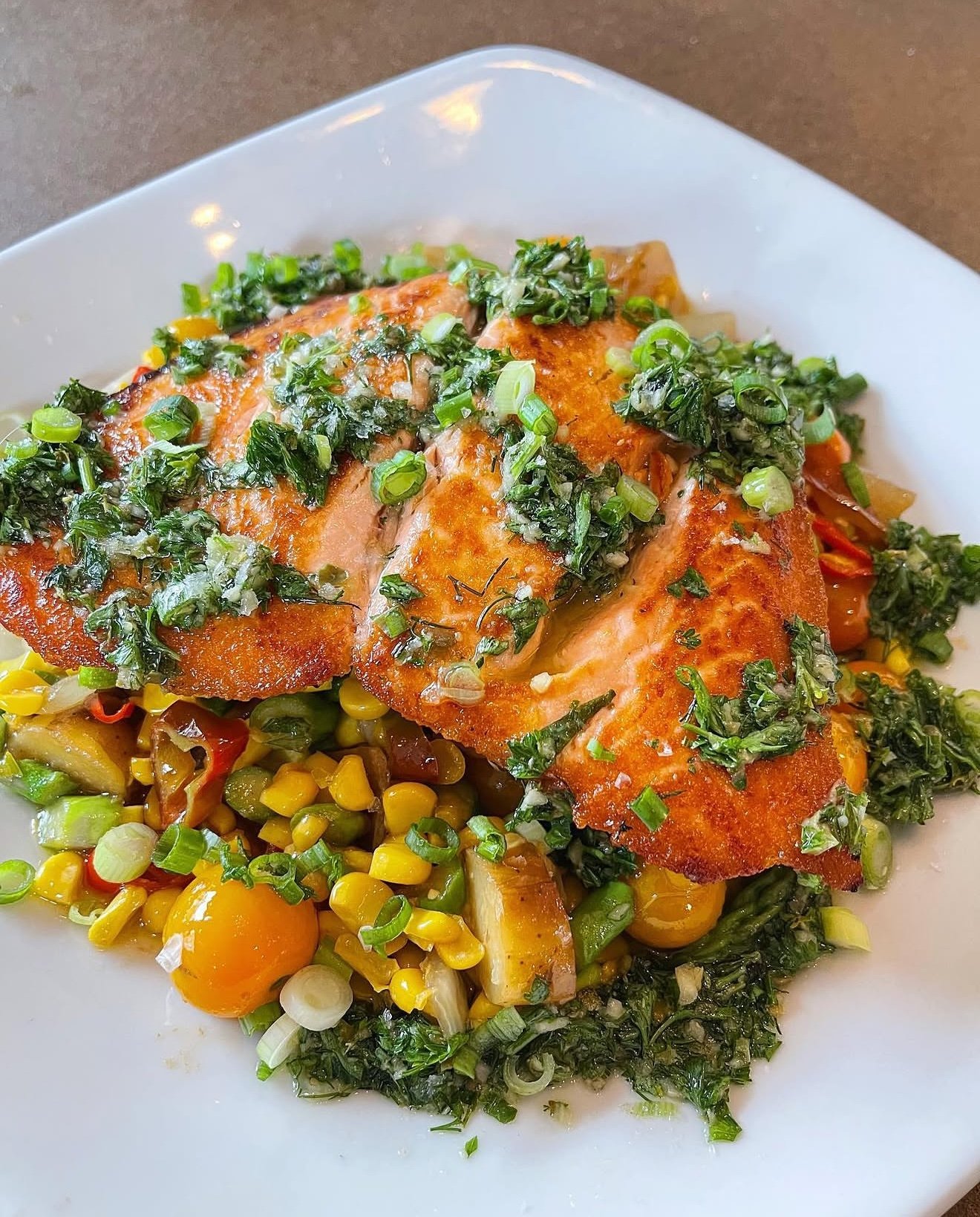 Pan-seared salmon topped with chopped herbs, served with a side of mixed vegetables including corn, cherry tomatoes, and green onions.