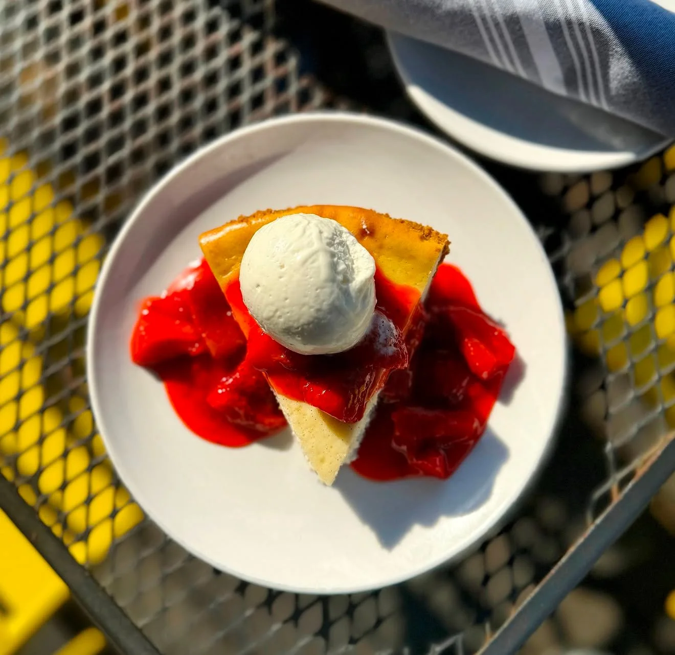 A plate of cheesecake topped with a scoop of vanilla ice cream, with sliced strawberries and strawberry sauce, placed on a yellow and gray mesh surface.