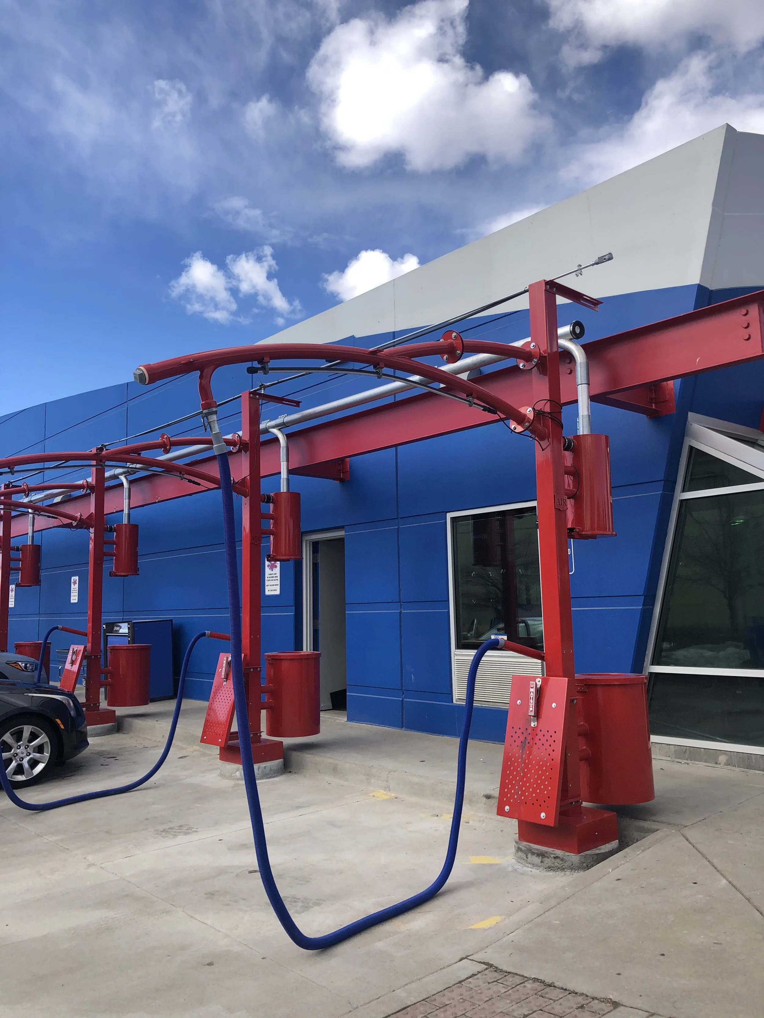 Self-serve car wash station with red equipment, blue hoses, and a blue building in the background, under a partly cloudy sky.